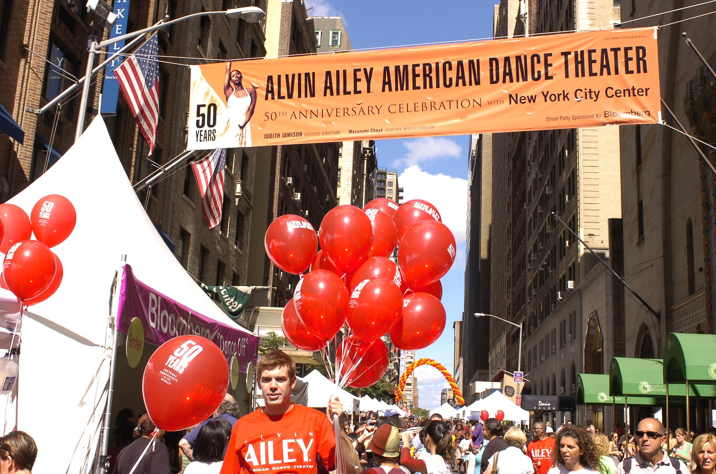 Street celebration for Alvin Ailey American Dance Theater's 50th Anniversary. A large banner reads Alvin Ailey American Dance Theater 50th Anniversary Celebration with New York City Center. People are gathered, holding red balloons with the 50 years logo. American flags and a white tent are visible. 