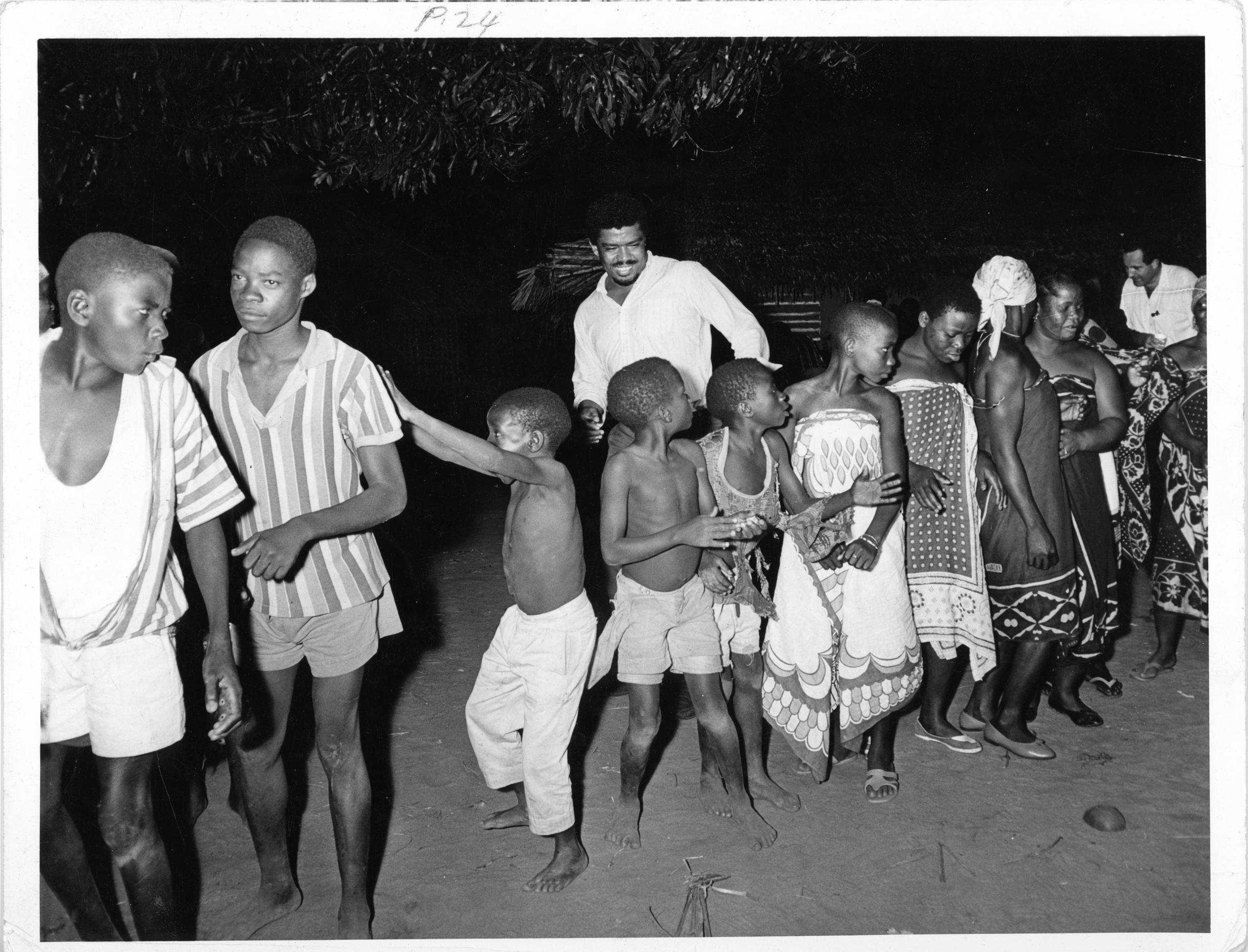 Black and white photo of Alvin Ailey with women and children of the Makonde Tribe in Africa, 1967. Ailey is smiling, standing among a group of people engaged in a line dance. The group includes both children and adults, wearing traditional clothing. 