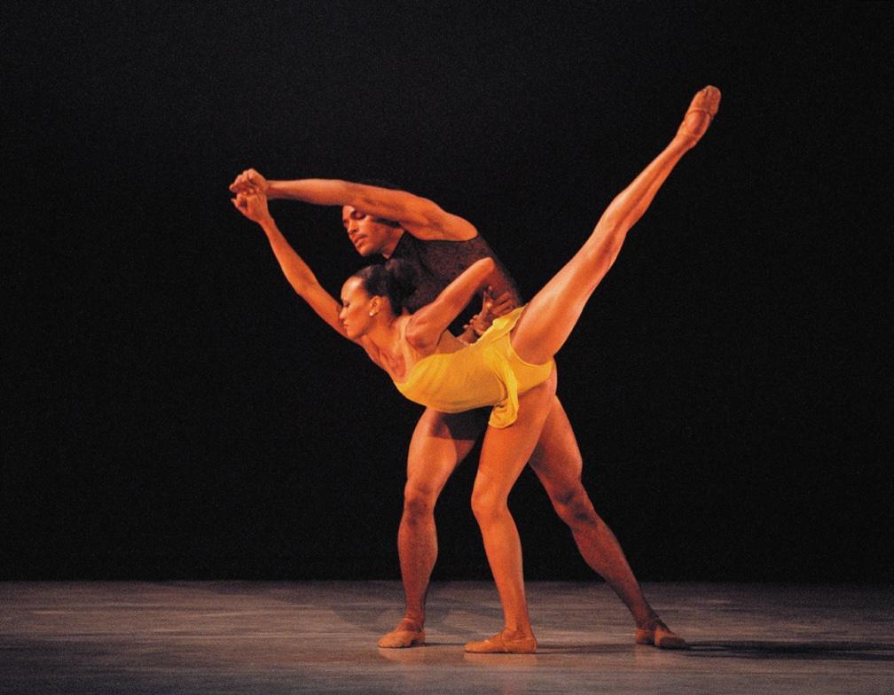 Two dancers from Alvin Ailey American Dance Theater performing. The male dancer supports the female dancer, who is wearing a yellow outfit and extending her leg high in an elegant pose. The background is dark, emphasizing the dancers.