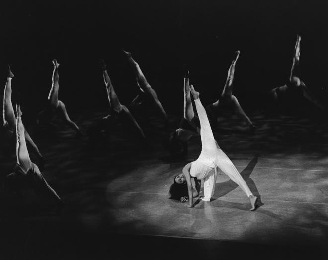 Black and white photo of Donna Wood and company members performing in Judith Jamison's "Divining." Wood is in the foreground, extending one leg upward in an elegant pose, while other dancers in the background mirror similar movements. The stage lighting casts dramatic shadows. Photo by Jack L. Vartoogian, 1984.