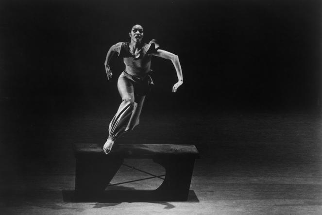 Black and white photo of Donna Wood performing in Ulysses Dove's "Inside." Wood is captured mid-movement, leaping over a bench with one foot on it. Her arms are positioned expressively, and she appears focused. The stage background is dark, highlighting her figure. Photo by William Hilton, 1979.