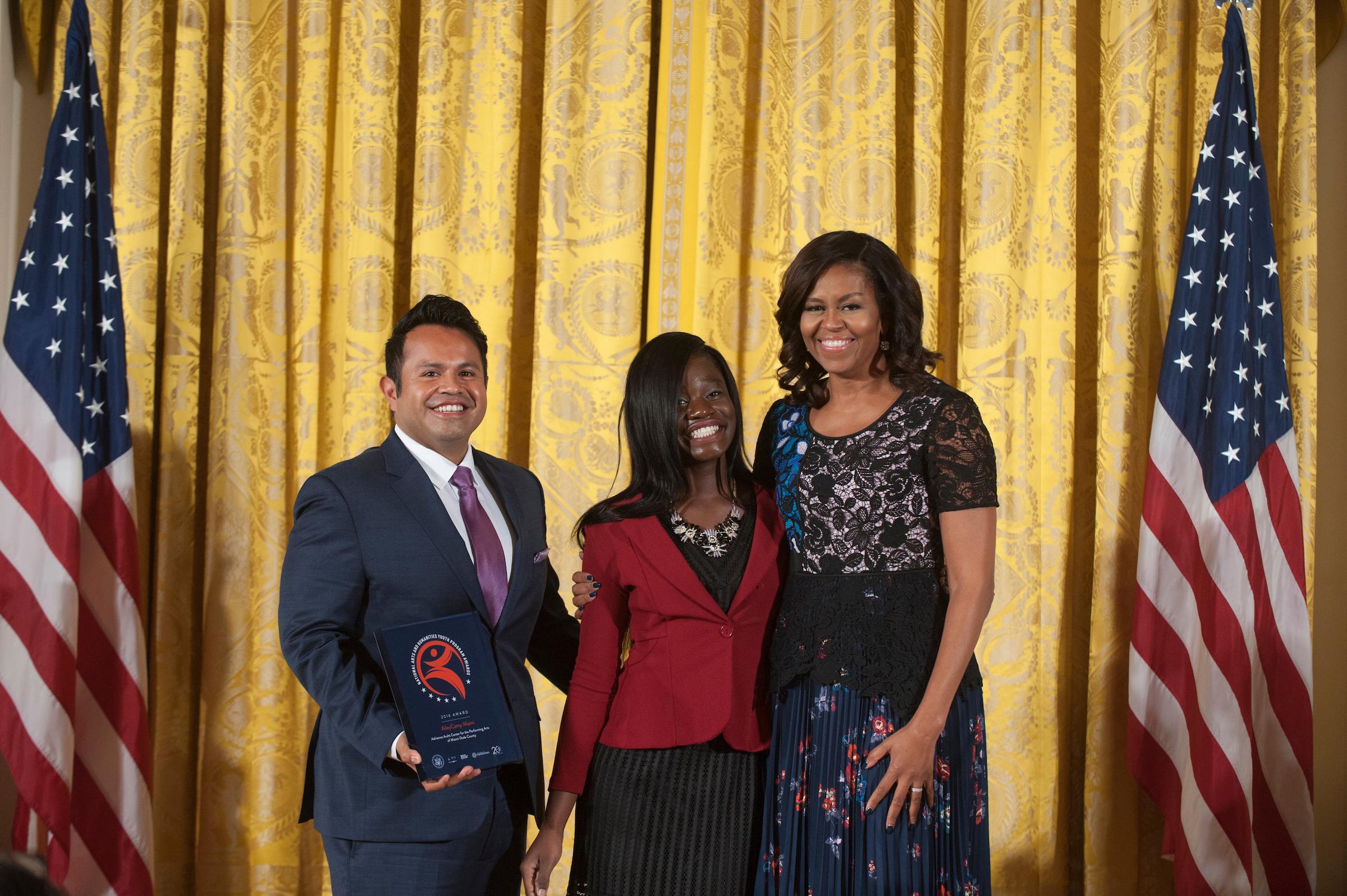 Jairo Ontiveros, AileyCamper Janasia Johnson, and Michelle Obama standing together in front of a gold curtain with American flags on either side. Ontiveros holds an award plaque. All three are smiling. Photo by Steven Purcell, 2016.