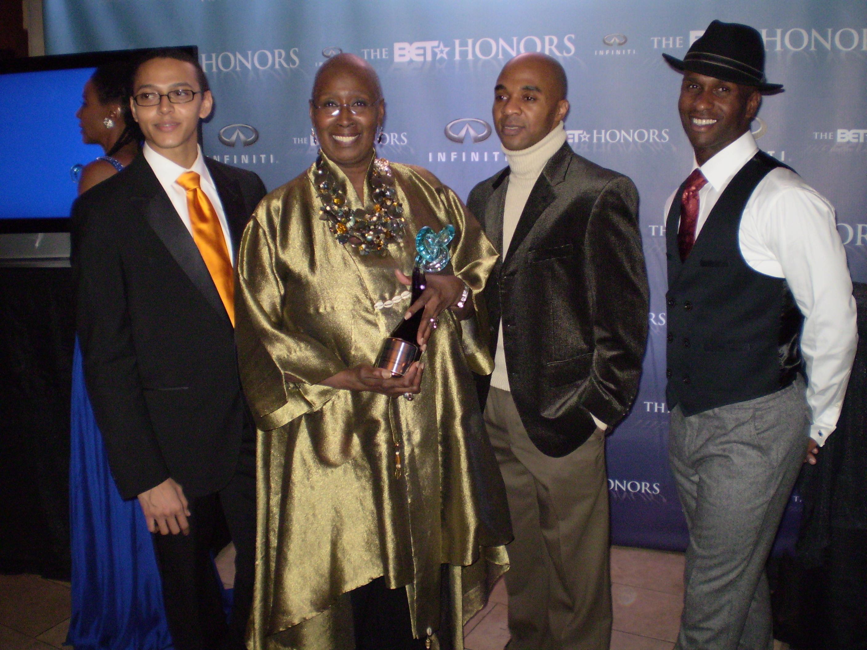 Judith Jamison and Alvin Ailey American Dance Theater’s Clifton Brown, Matthew Rushing, and Glenn Allen Sims at the BET Honors in January 2009. Jamison is holding an award and dressed in a gold outfit, flanked by Brown, Rushing, and Sims, who are all smiling and dressed in formal attire. The backdrop features the BET Honors logo. Photo by Christopher Zunner, 2009.