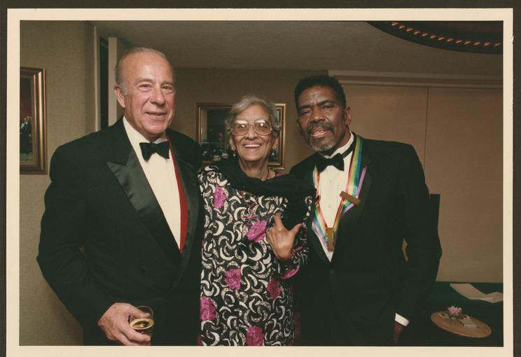  In this photograph from 1988, Alvin Ailey is pictured at the Kennedy Center Honors alongside Lula Cooper and George Schultz, the U.S. Secretary of State. Alvin Ailey, adorned with the colorful ribbon of the Kennedy Center Honors medal, is smiling warmly, symbolizing his recognition and achievements in the arts. Lula Cooper, elegantly dressed in a floral patterned outfit, stands between them, exuding grace. George Schultz is on the left, dressed in a classic black tuxedo, holding a drink and smiling. The se