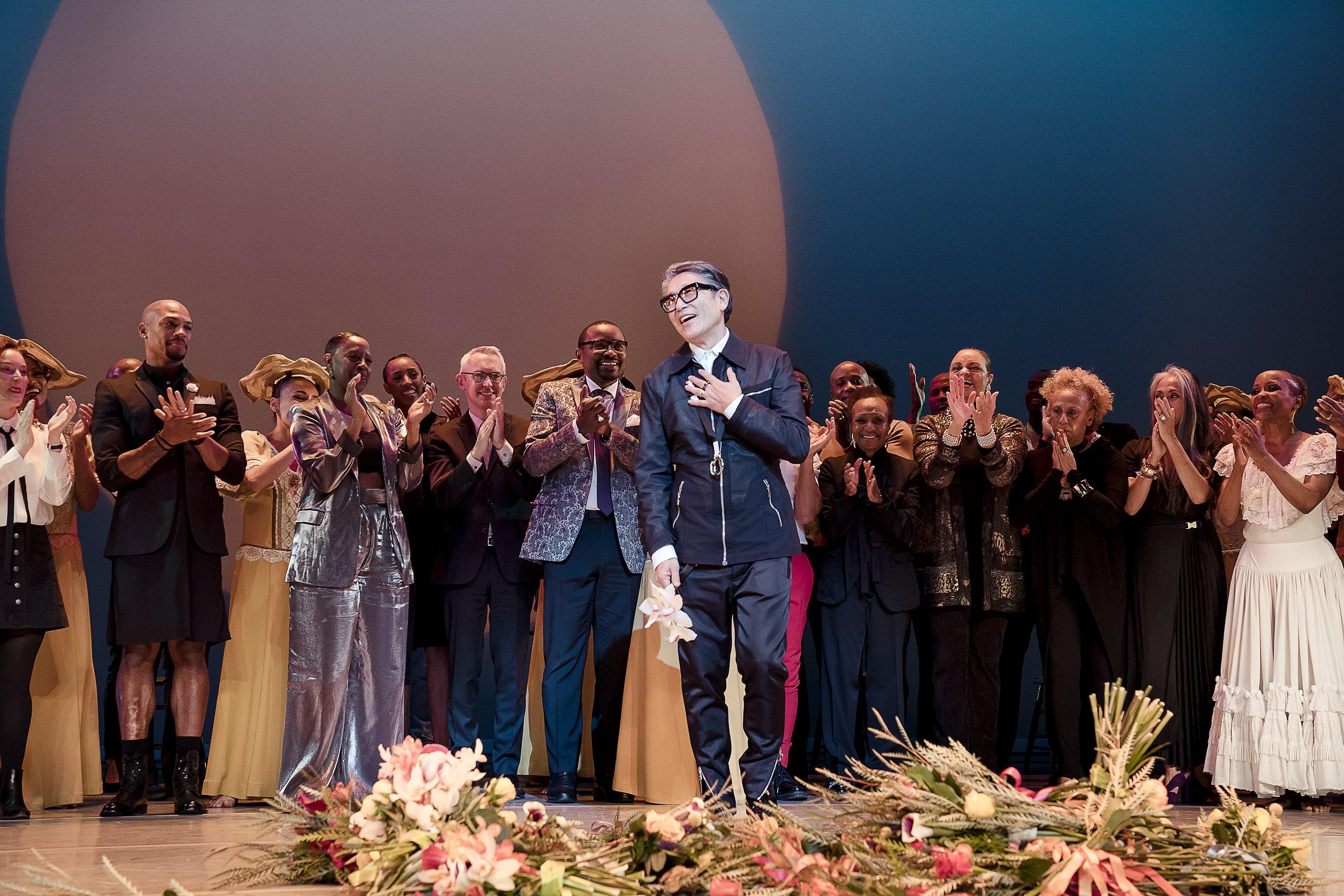 In this image from December 22, 2019, Masazumi Chaya is being applauded on stage at New York City Center after the "Celebrating Chaya" tribute program. The photo captures a moment of heartfelt recognition and appreciation for Chaya's contributions. Chaya stands at the center, holding a bouquet of flowers, with his hand on his heart, a gesture of gratitude. Surrounding him are dancers, colleagues, and admirers, all clapping and smiling in admiration. The stage is adorned with floral arrangements, and the lig