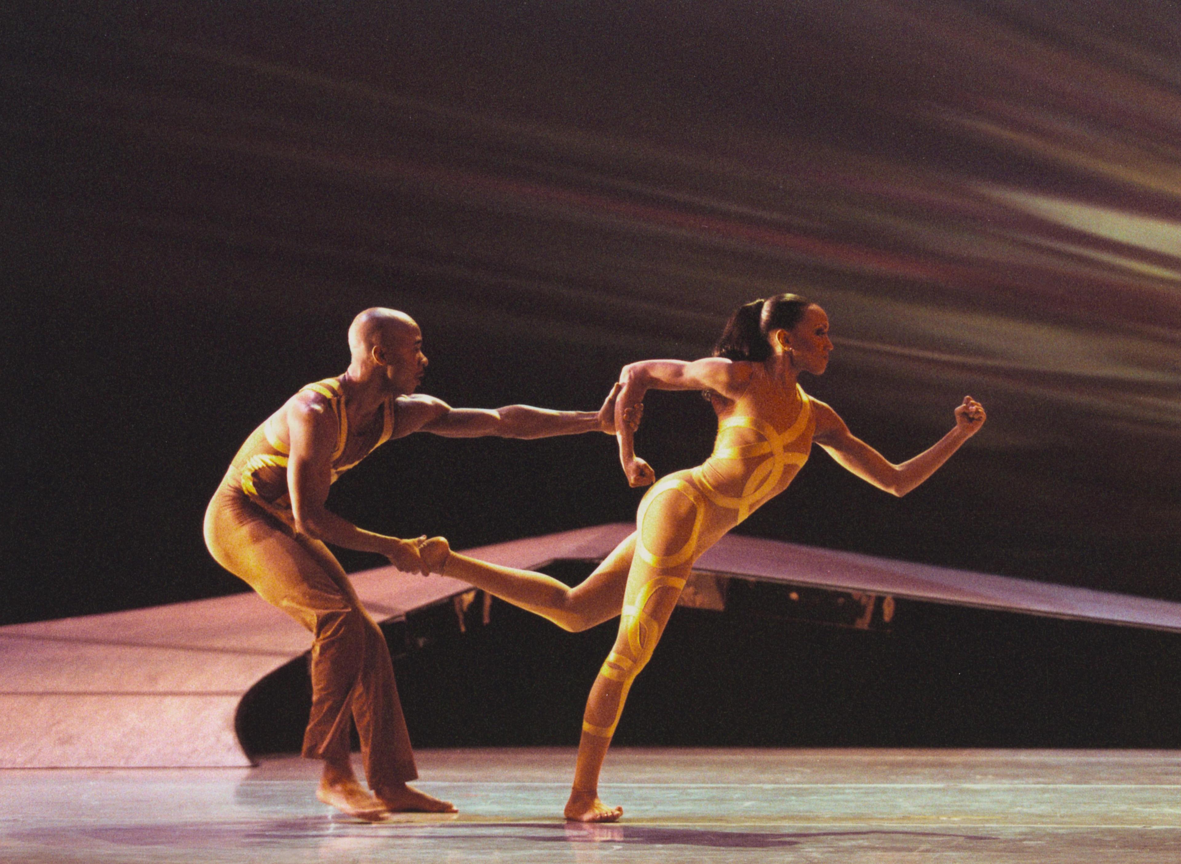  This vibrant photograph captures Matthew Rushing and Linda-Denise Fisher-Harrell performing in "HERE...NOW," a dance piece. The image, taken by Paul Kolnik in 2001, showcases the dynamic energy and physicality of the performance. Rushing is seen gripping Fisher-Harrell's hand as she leans forward, both dancers adorned in sleek, form-fitting costumes that emphasize their movements. The background features a blurred, abstract set design that enhances the sense of motion and intensity. This moment reflects th