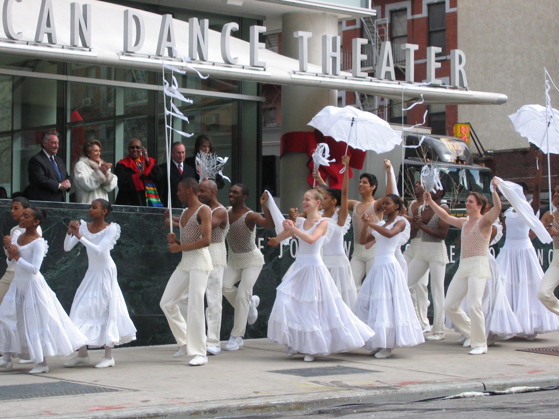  This image captures the ribbon-cutting ceremony at the opening of the Joan Weill Center for Dance in 2005. The celebratory moment features dancers in white attire performing outside the building, symbolizing the joyous occasion. Notable figures, including dignitaries and guests, are visible in the background, adding to the event's significance. The dancers' coordinated movements, along with the white flags and umbrellas, reflect a sense of unity and celebration, marking a pivotal moment in the history of t