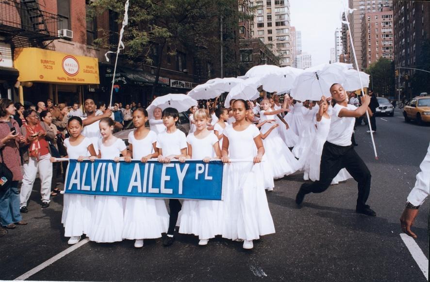 This image captures a joyful parade featuring students from The Ailey School marching to the future site of Alvin Ailey American Dance Theater's new home. The students are holding a banner that reads "ALVIN AILEY PL," symbolizing the significance of this momentous occasion. Dressed in white and carrying umbrellas, the students are accompanied by a performer dancing energetically alongside them. The parade took place in 2002 and was documented by photographer Kwame Brathwaite. This event marks a pivotal mome