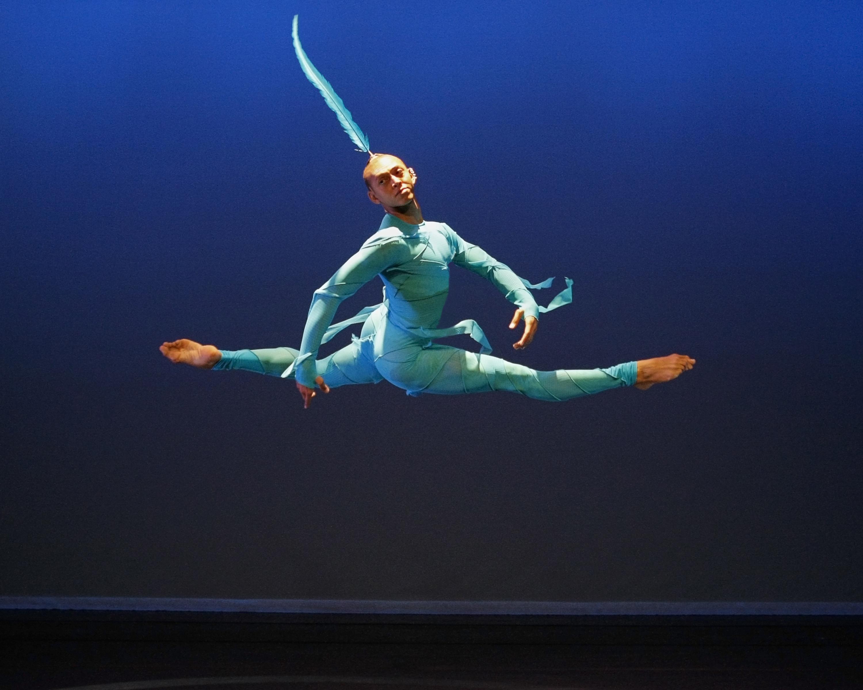 Man with split legs in the air wearing a long single feather from his bald head