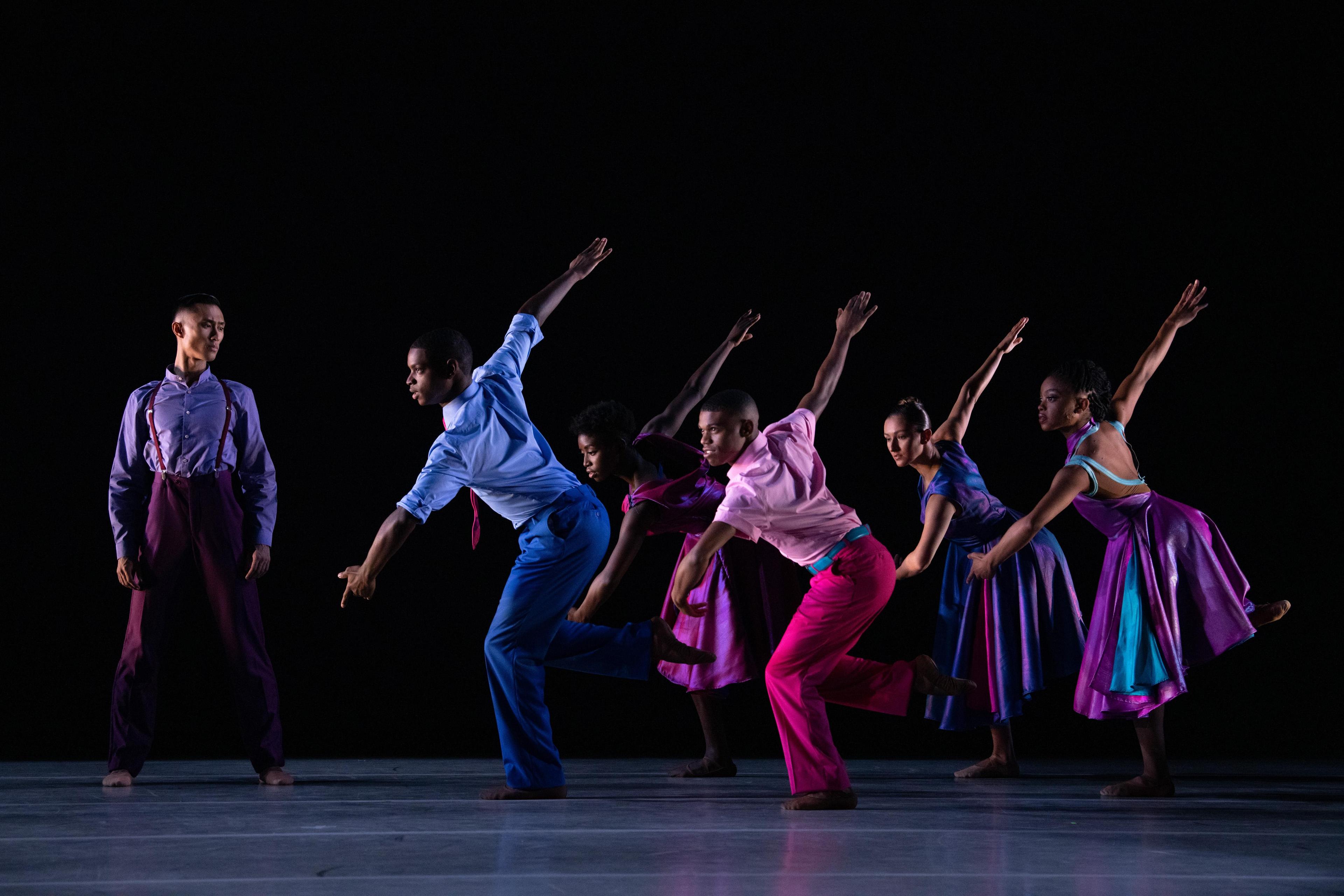 A group of dancers in colorful costumes perform on stage against a dark background. They are in a dynamic formation, with arms extended and bodies leaning forward, showcasing movement and balance.