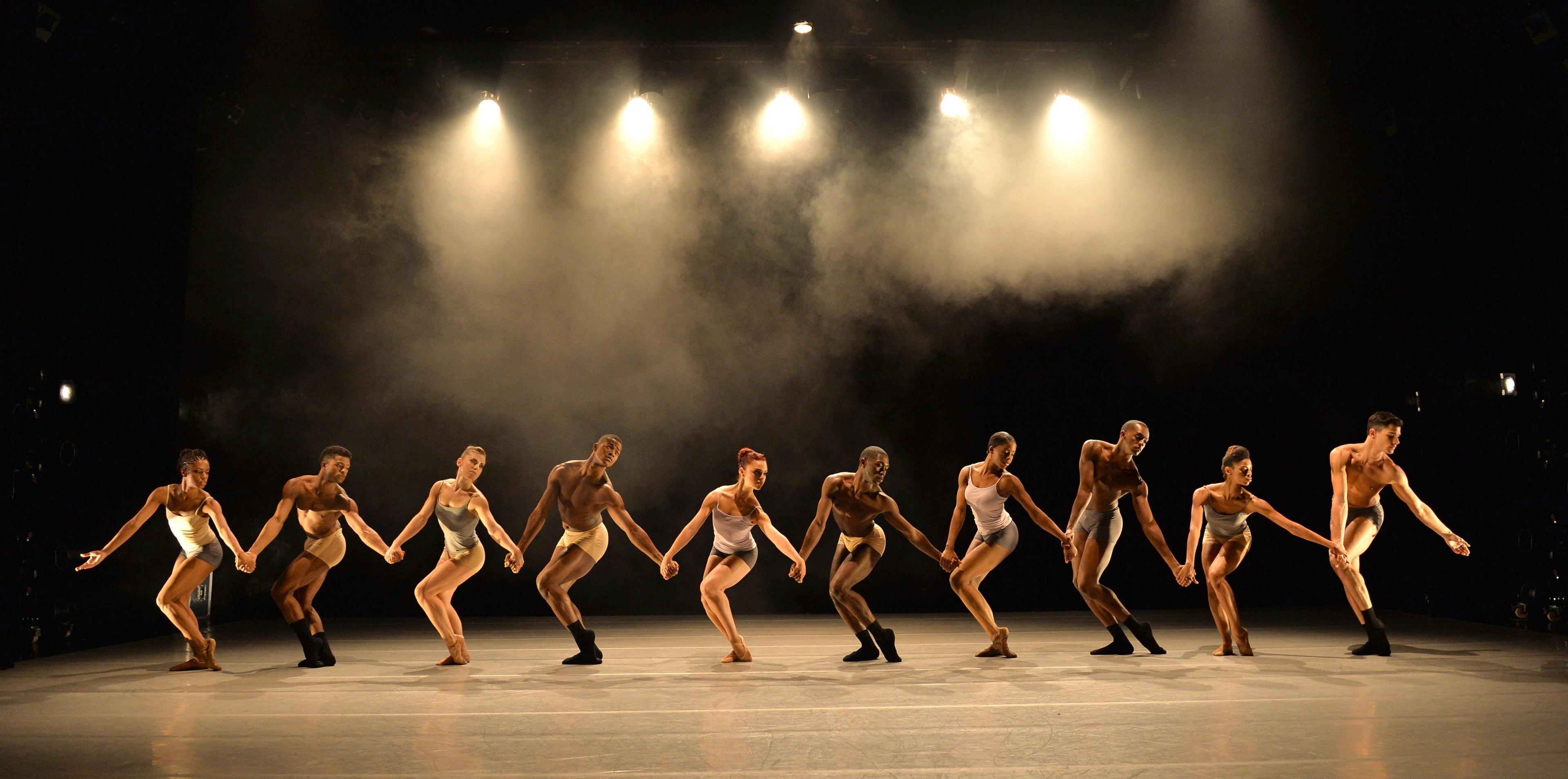 A group of dancers in minimal, neutral-toned costumes perform in a line formation on stage, holding hands. They are illuminated by stage lights against a smoky, dark background, creating a dramatic atmosphere.