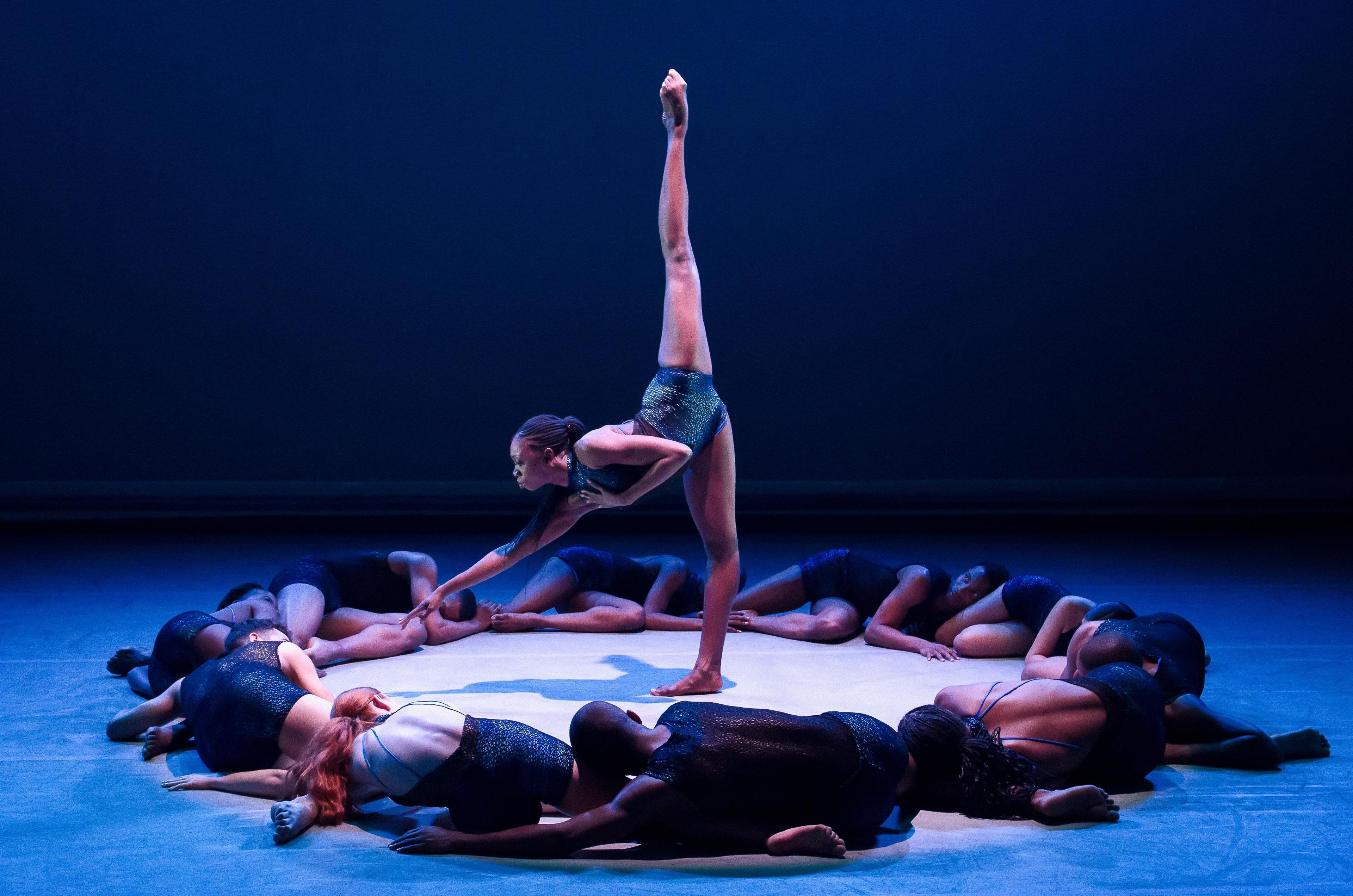 A group of dancers is arranged in a circle on the stage, lying down with their heads towards the center. One dancer stands in the middle, balancing on one leg with the other leg extended high in the air. They are dressed in dark, sparkly costumes, and the stage is lit with blue lighting.