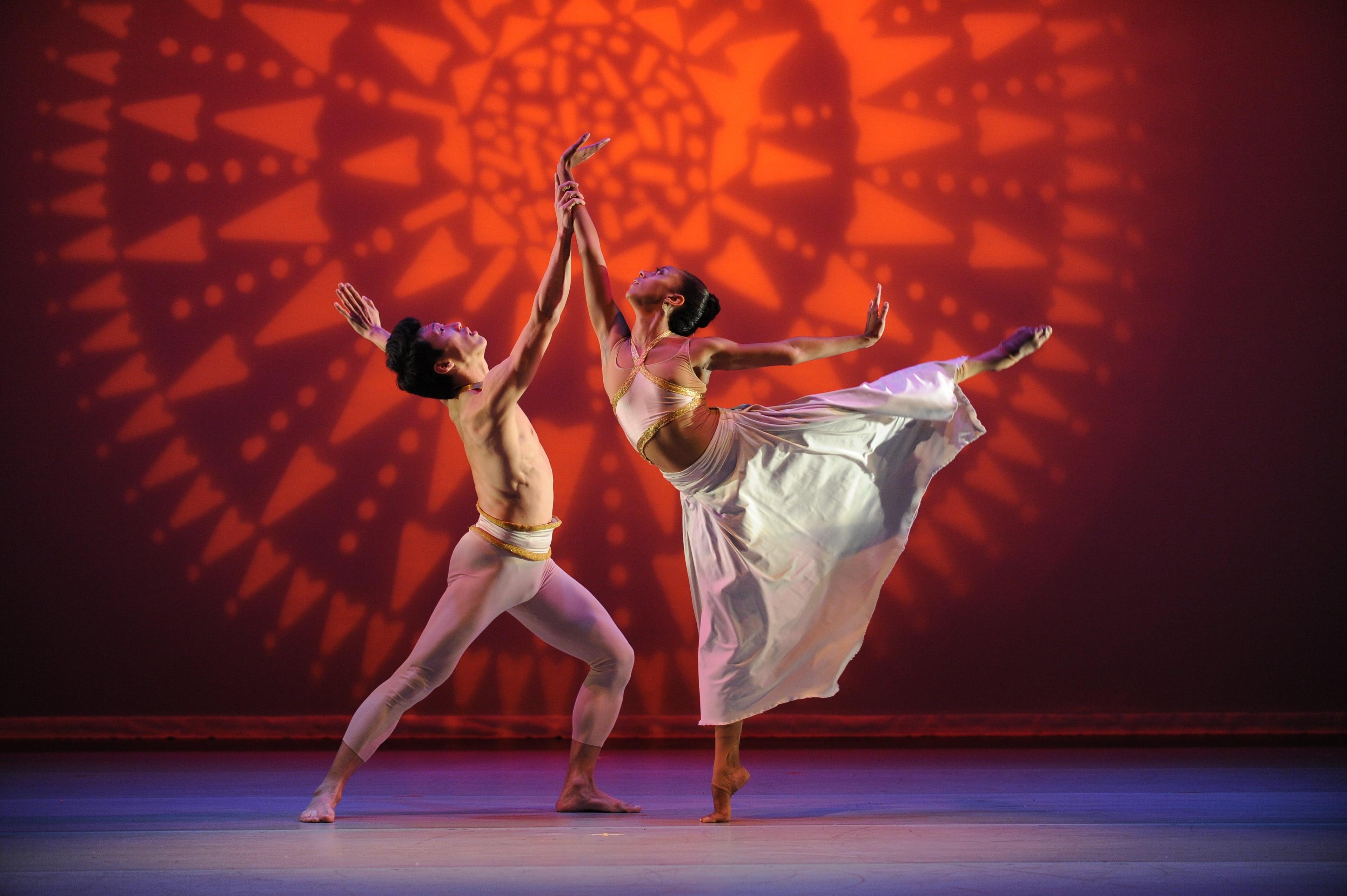 Two dancers are performing a duet on stage. The male dancer is in a lunge position, wearing beige tights with gold accents. The female dancer is balancing on one leg with the other leg extended behind her, wearing a flowing white skirt and gold top. They are holding hands and reaching upward, with an intricate orange and red patterned background.