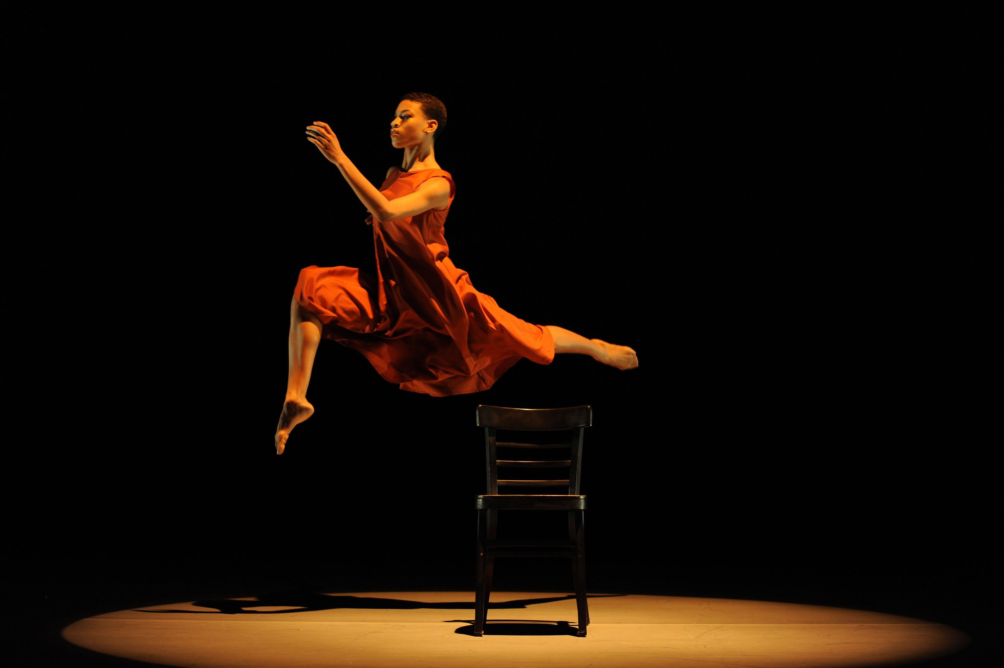 A dancer in an orange dress is captured mid-leap over a wooden chair on stage. The background is dark, and a spotlight illuminates the dancer and the chair, emphasizing the grace and height of the jump.