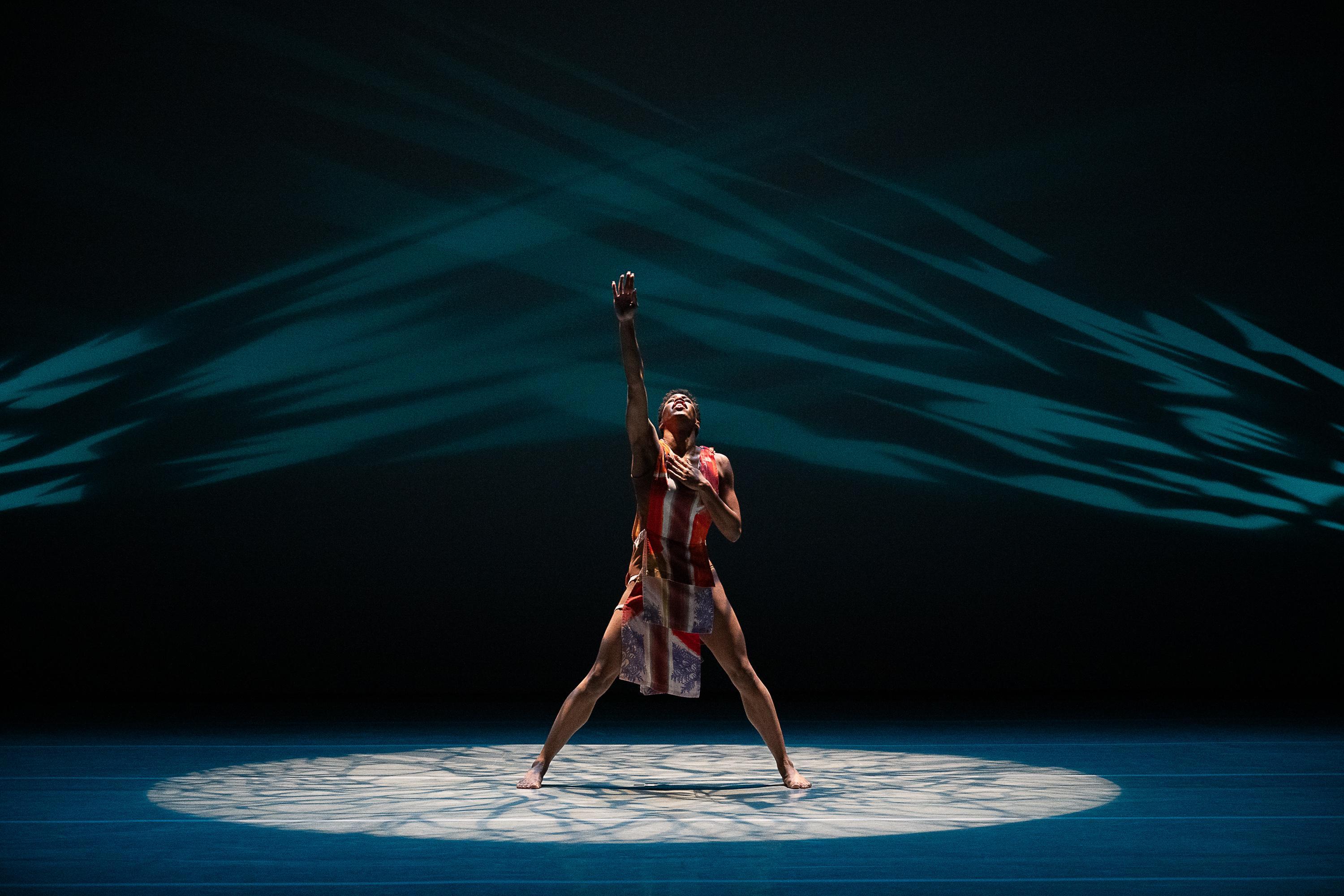 A dancer is performing on stage, standing in a wide stance with one arm reaching upward and the other hand on their chest. They are wearing a colorful, patterned costume. The stage is lit with a spotlight on the dancer, and the background features dynamic blue and black lighting patterns.