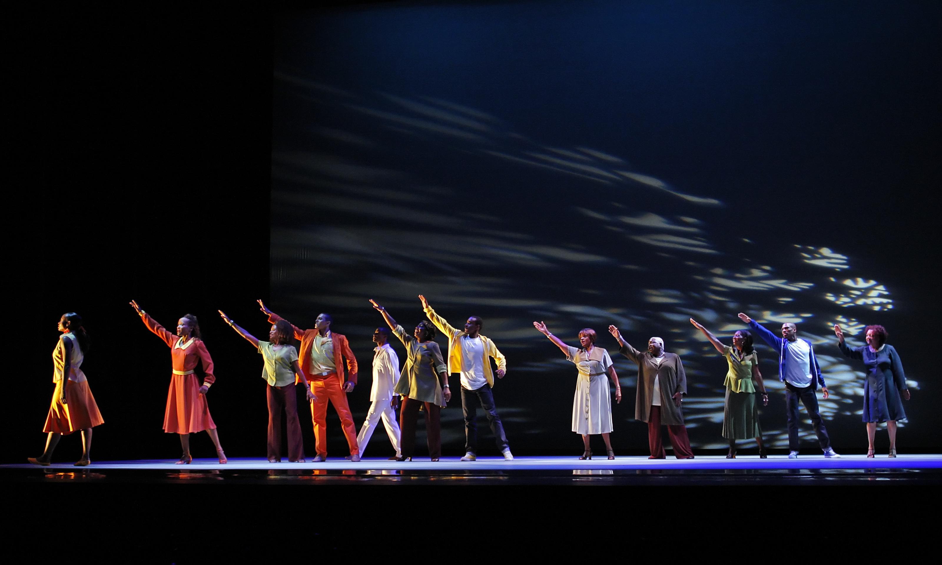 A group of dancers in colorful outfits standing in a line on stage, each with one arm extended upward. The background is dark with patterns of light casting shadows, creating a dynamic visual effect. The dancers wear various shades of orange, yellow, green, and blue.