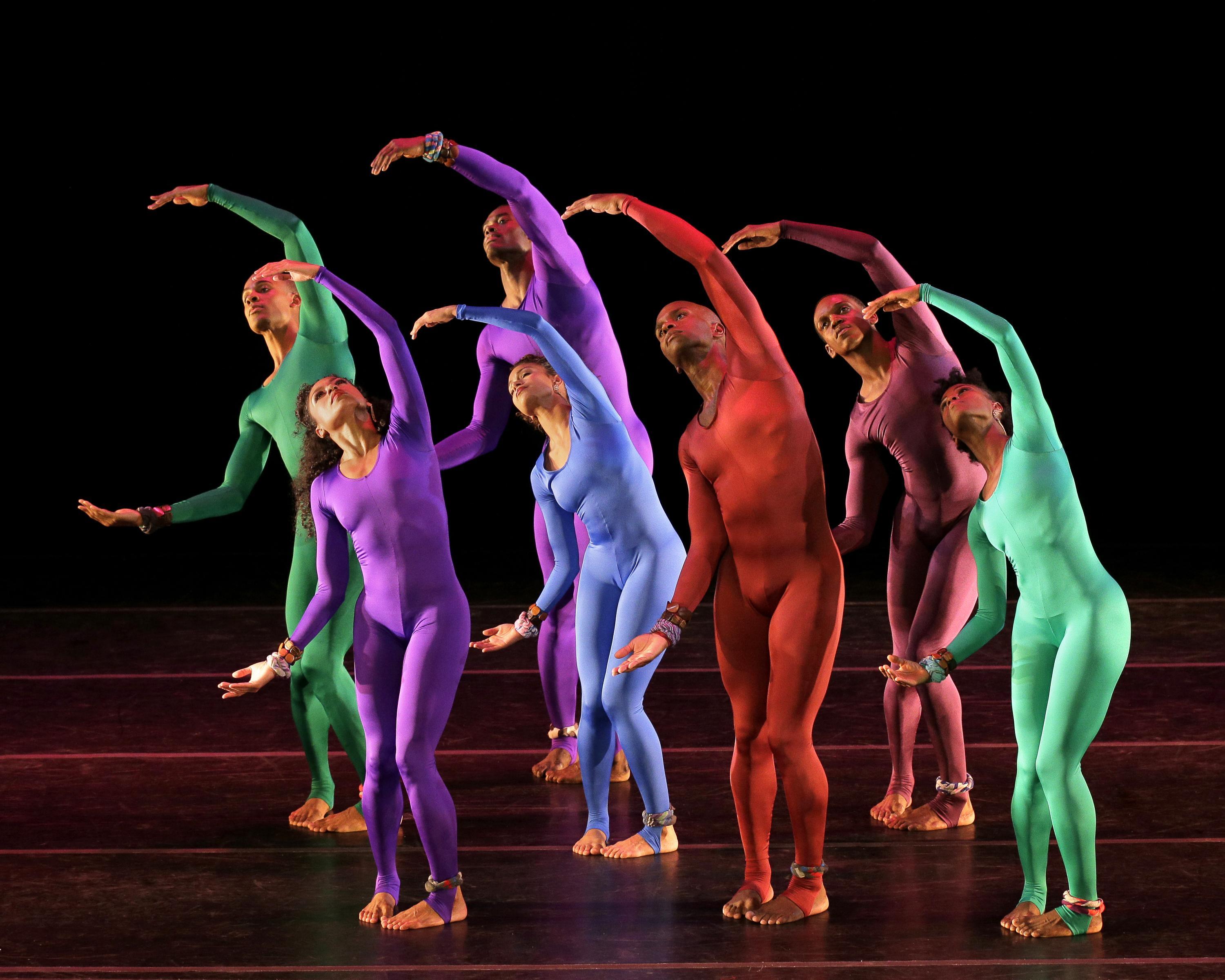 Dancers in brightly colored bodysuits (green, purple, blue, red) performing on stage, forming a wave-like pattern with their arms. The background is dark, making their vibrant outfits stand out. The dancers' movements are synchronized, creating a visually captivating and fluid scene.