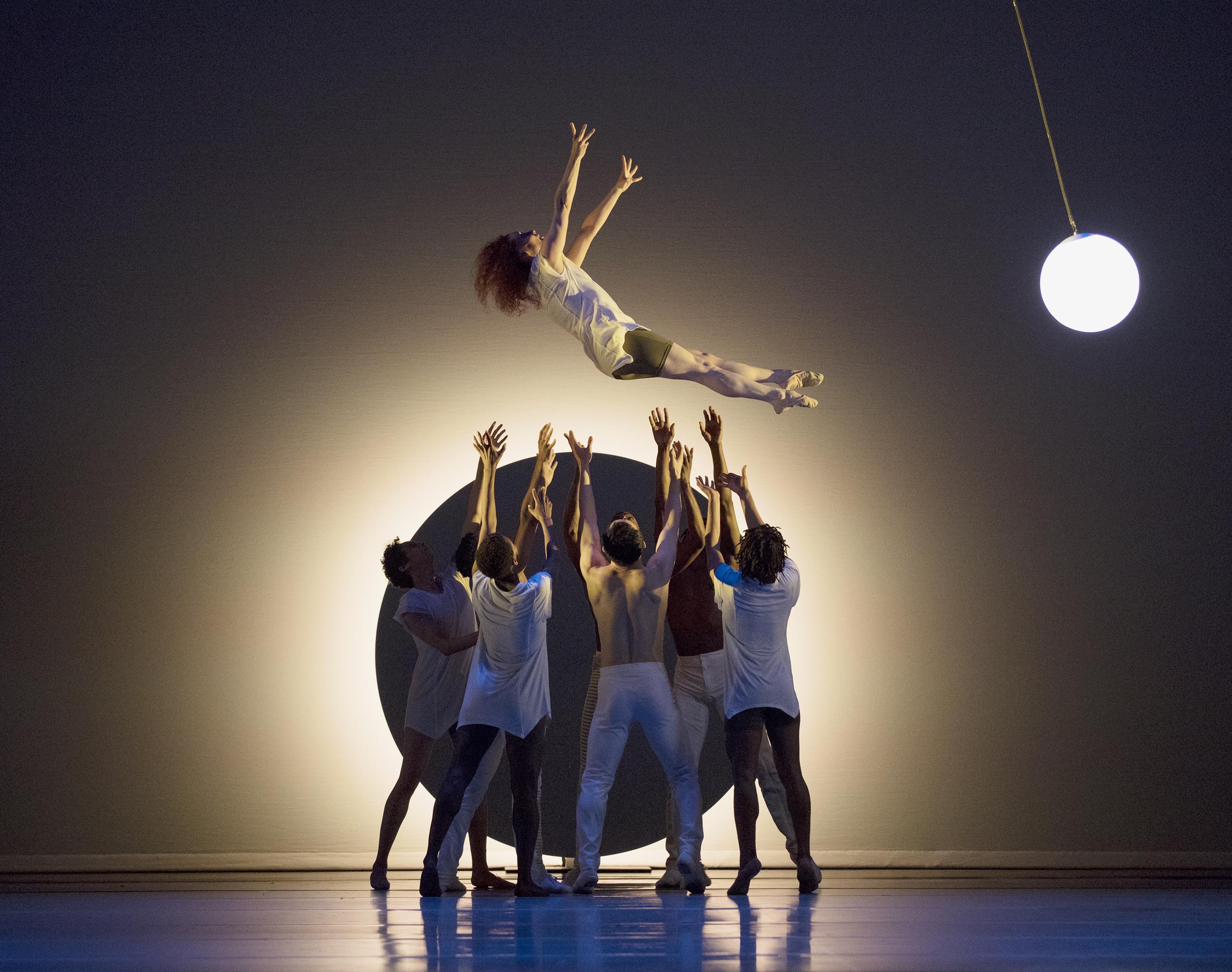 Dancers on stage, with one dancer being lifted high in the air by a group of others. The background features a large, glowing circular light and a hanging spherical lamp. The dancers are dressed in light-colored costumes, creating a dramatic and ethereal scene.