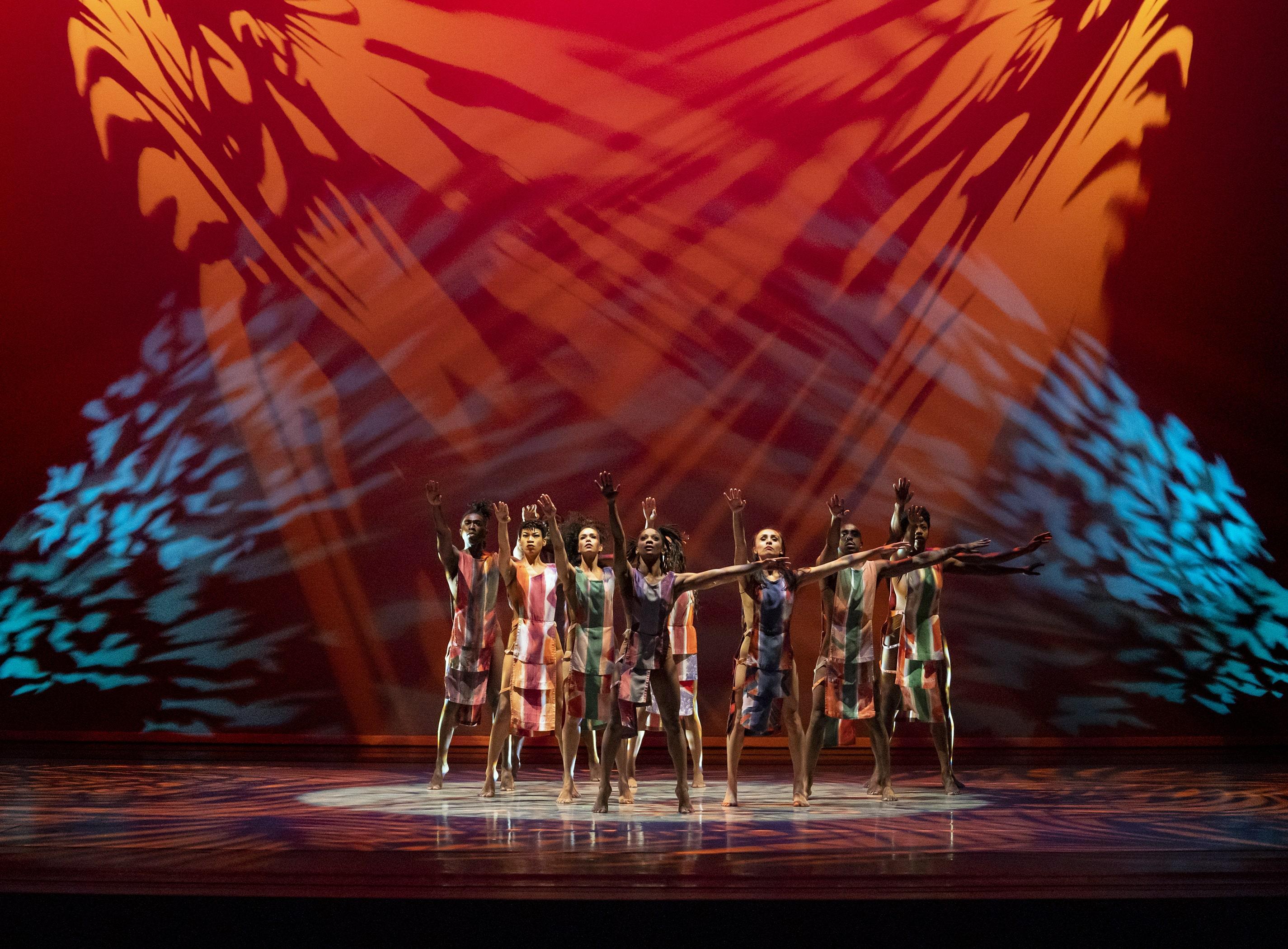 Dancers on stage in a vibrant performance, wearing multicolored costumes. They form a line with arms raised, creating a dynamic visual. The background is illuminated with dramatic red, orange, and blue patterns, enhancing the energetic and lively atmosphere of the scene.