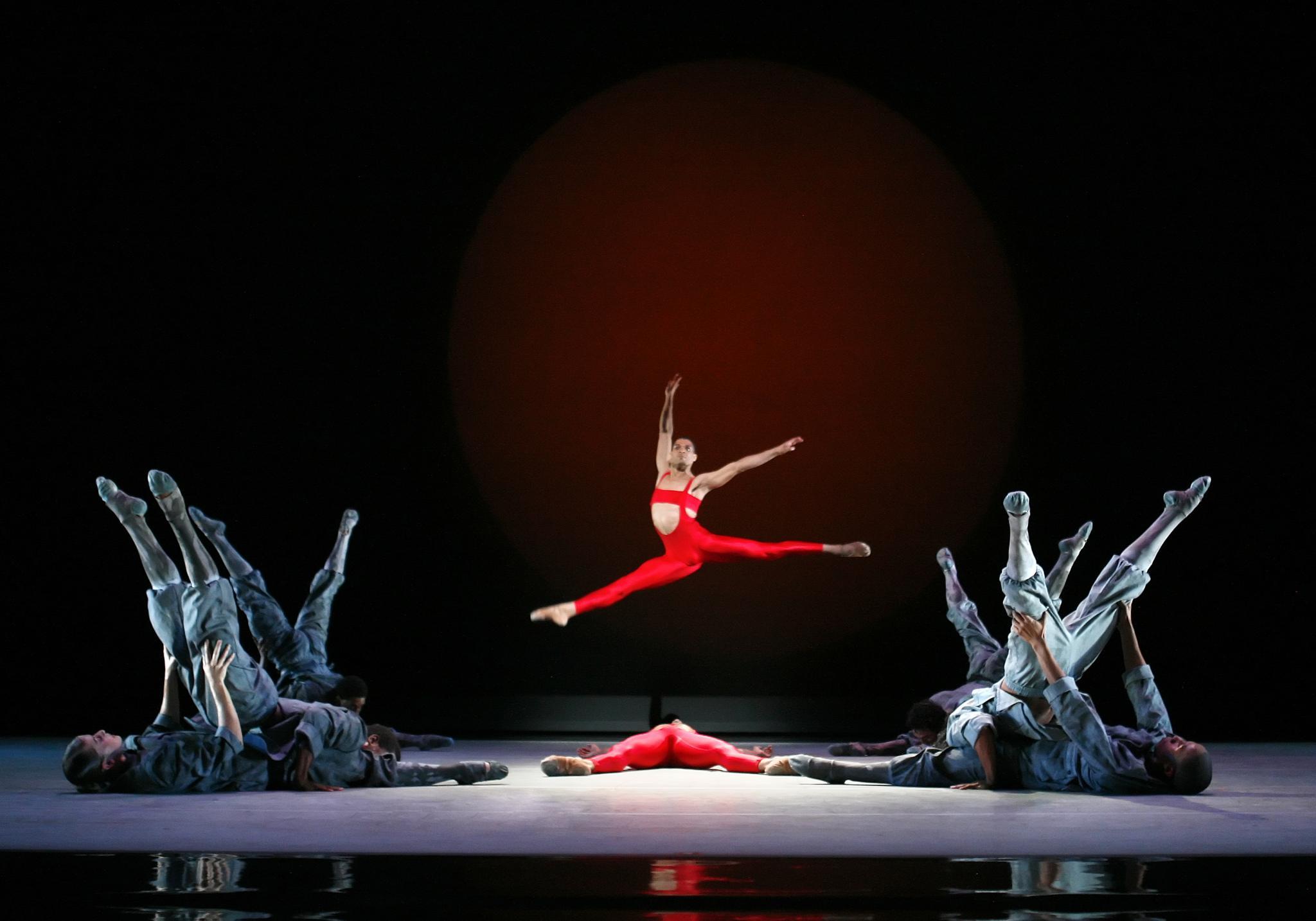 A dancer in a bright red costume performs a mid-air split leap, framed by a large, glowing red circle in the background. Surrounding the leaping dancer, other dancers in grey costumes lie on the stage floor with their legs raised, creating a striking contrast and dynamic composition.