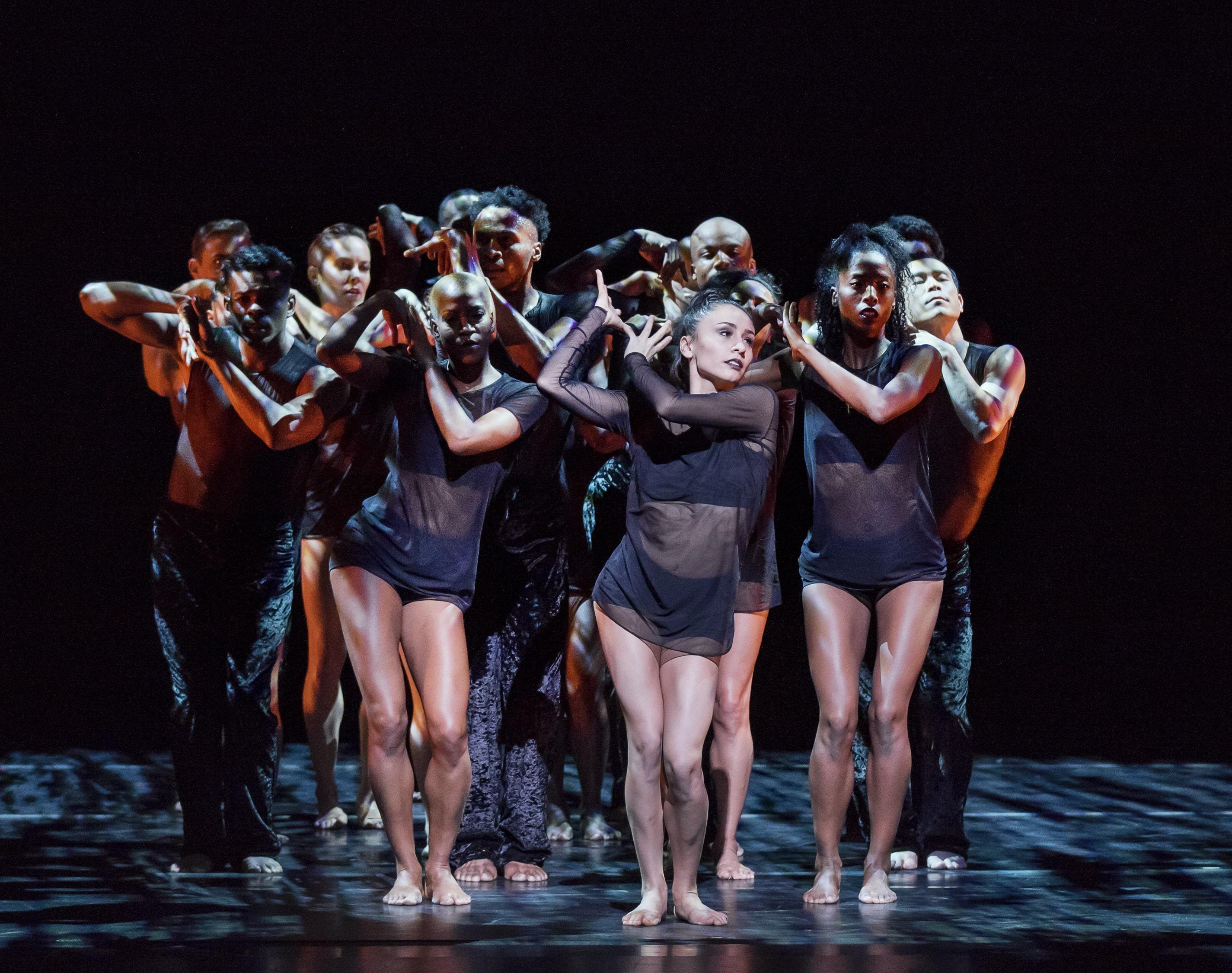 A group of dancers on stage, closely packed together, wearing black sheer outfits. They perform with intense expressions and synchronized arm movements, creating a powerful and unified formation. The background is dark, emphasizing their dramatic poses and the interplay of light and shadows on their bodies.