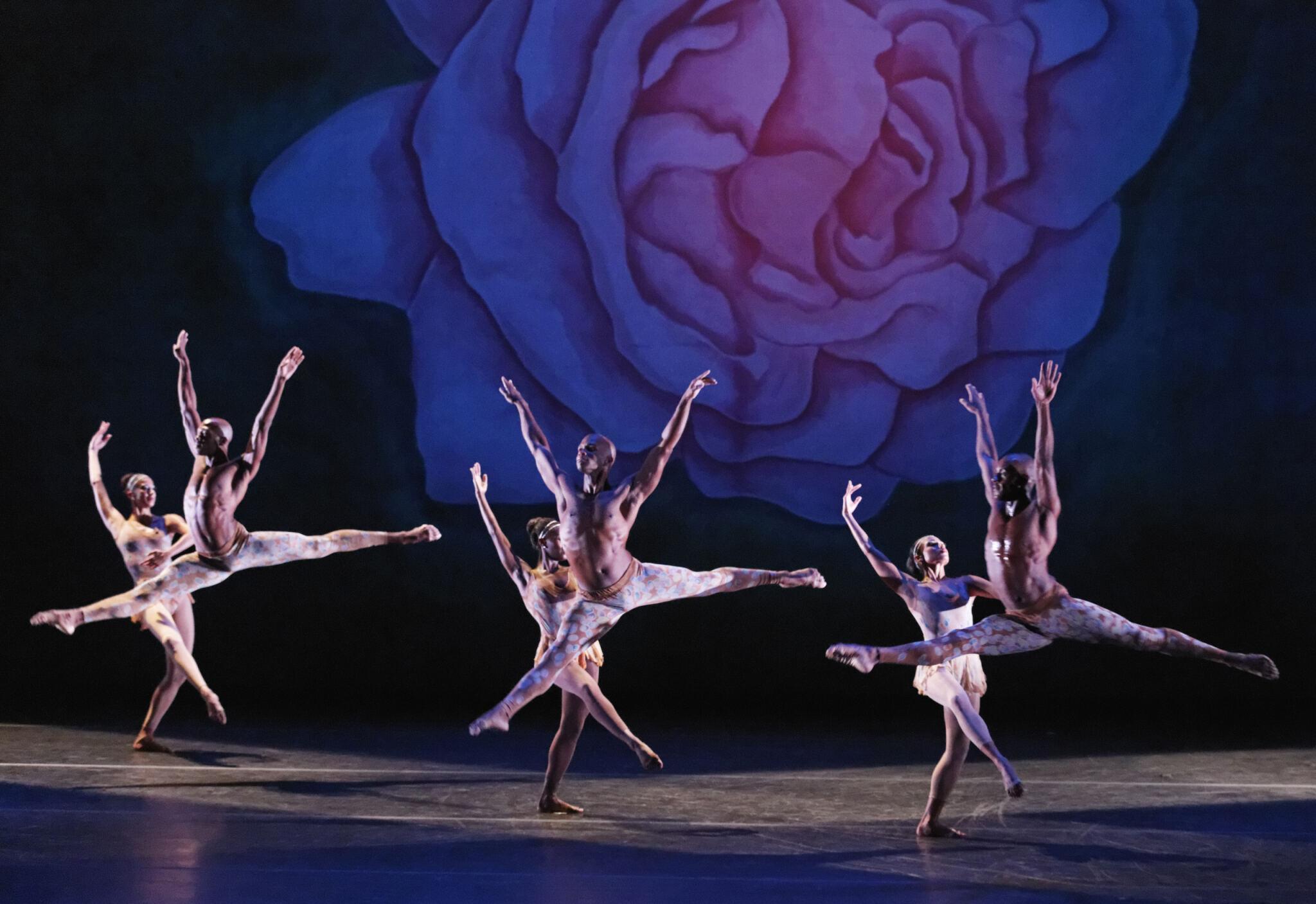 Dancers on stage performing dynamic leaps, wearing floral-patterned costumes. The backdrop features a large, detailed image of a pink and purple rose, enhancing the romantic and vibrant atmosphere of the scene. The dancers' movements are synchronized, creating a visually stunning and graceful performance.