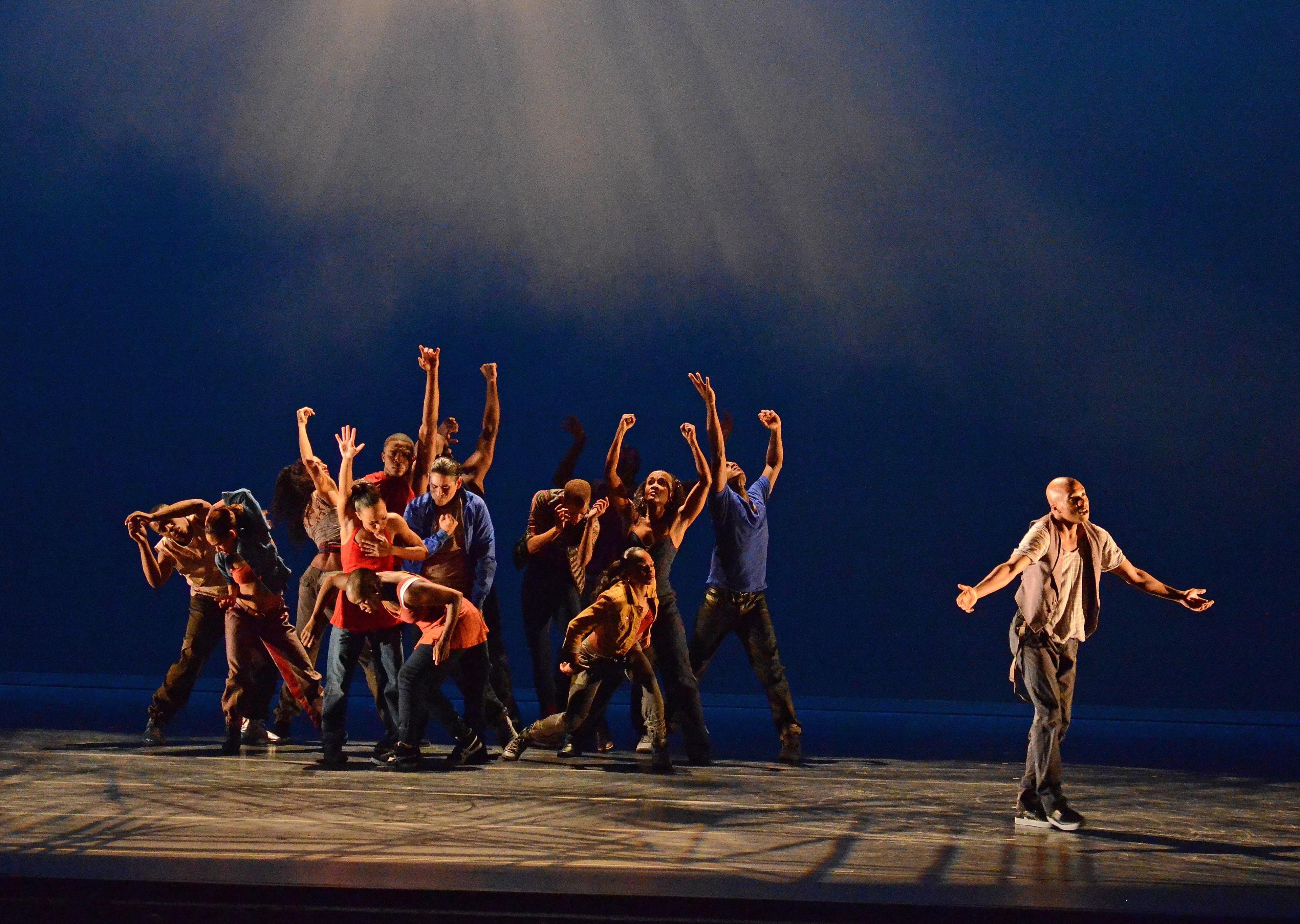 Dancers on stage in a dramatic formation, with one dancer standing apart from the group. They are dressed in casual clothing, and the lighting creates a spotlight effect on the ensemble. The group displays various expressive poses, conveying a sense of struggle and emotion. The background is dark blue, enhancing the intensity of the scene.