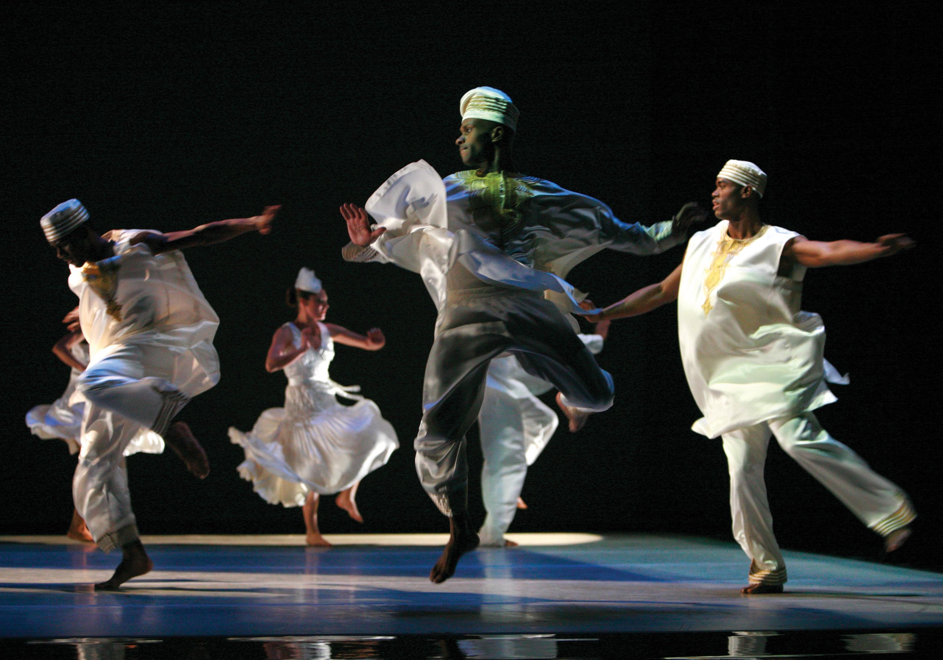 Dancers performing energetically on stage, dressed in traditional African-inspired white attire. Their movements are dynamic and expressive, with flowing garments accentuating their motions. The background is dark, focusing attention on the dancers' vibrant and powerful performance.