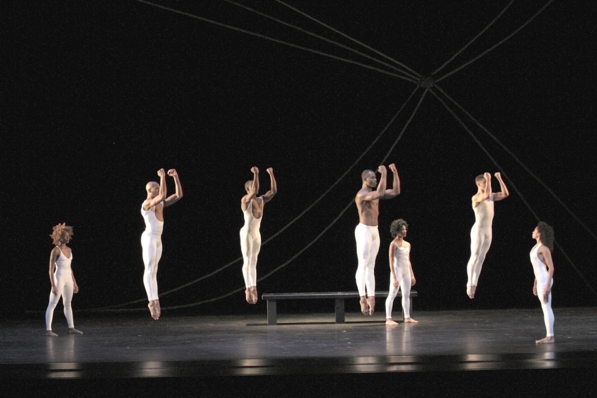 Dancers on stage in white costumes, captured mid-jump with arms bent at the elbows, creating a uniform, powerful visual. Some dancers stand on the ground, observing. A dark background with diagonal ropes adds a dramatic and structured element to the scene, emphasizing the height and precision of the jumps.