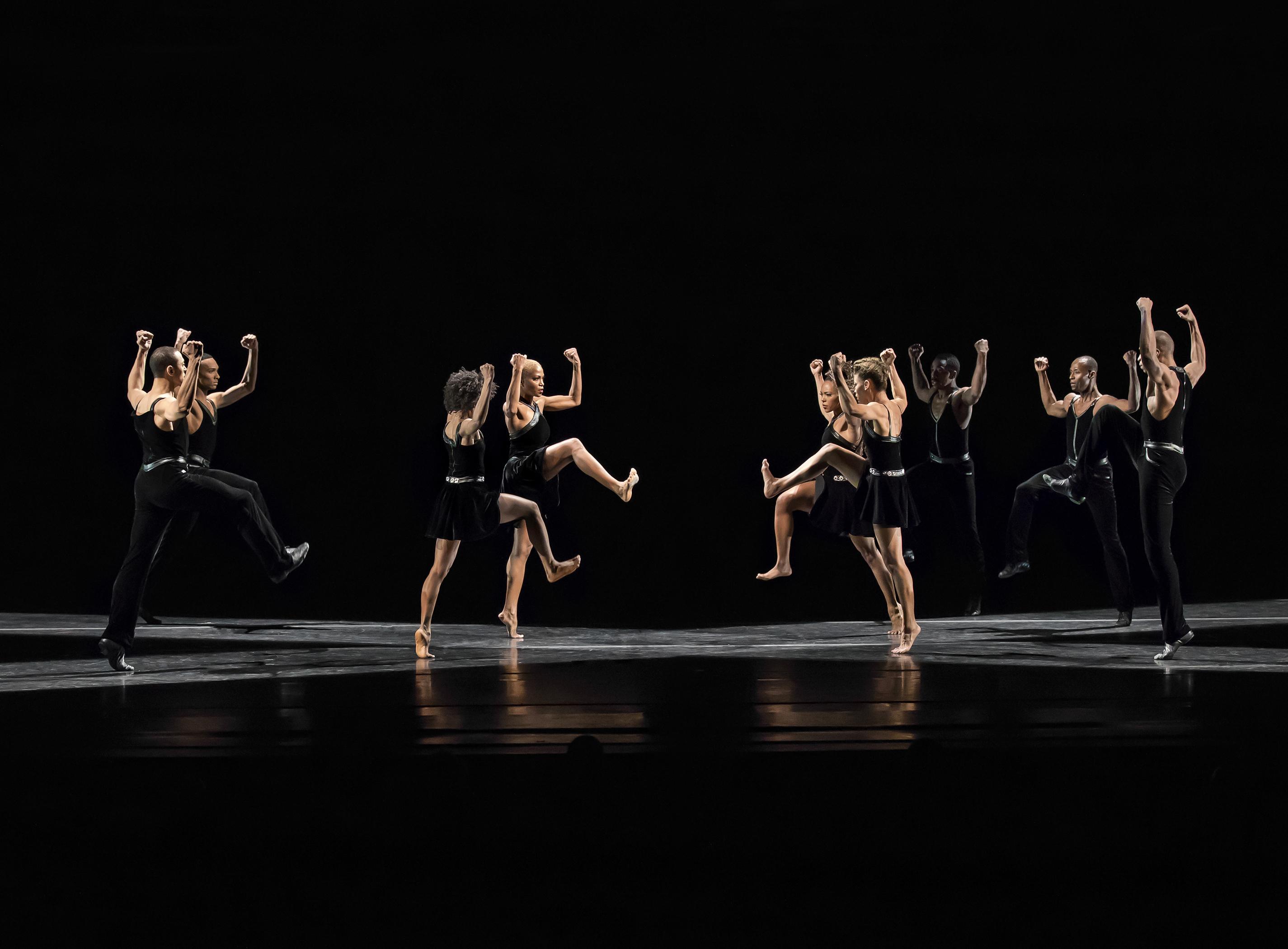 Dancers on stage in black costumes, performing in two groups facing each other. Their movements are synchronized, with raised arms and bent knees, creating a mirrored effect. The dark background highlights their strong, dynamic poses and the contrast between light and shadow.