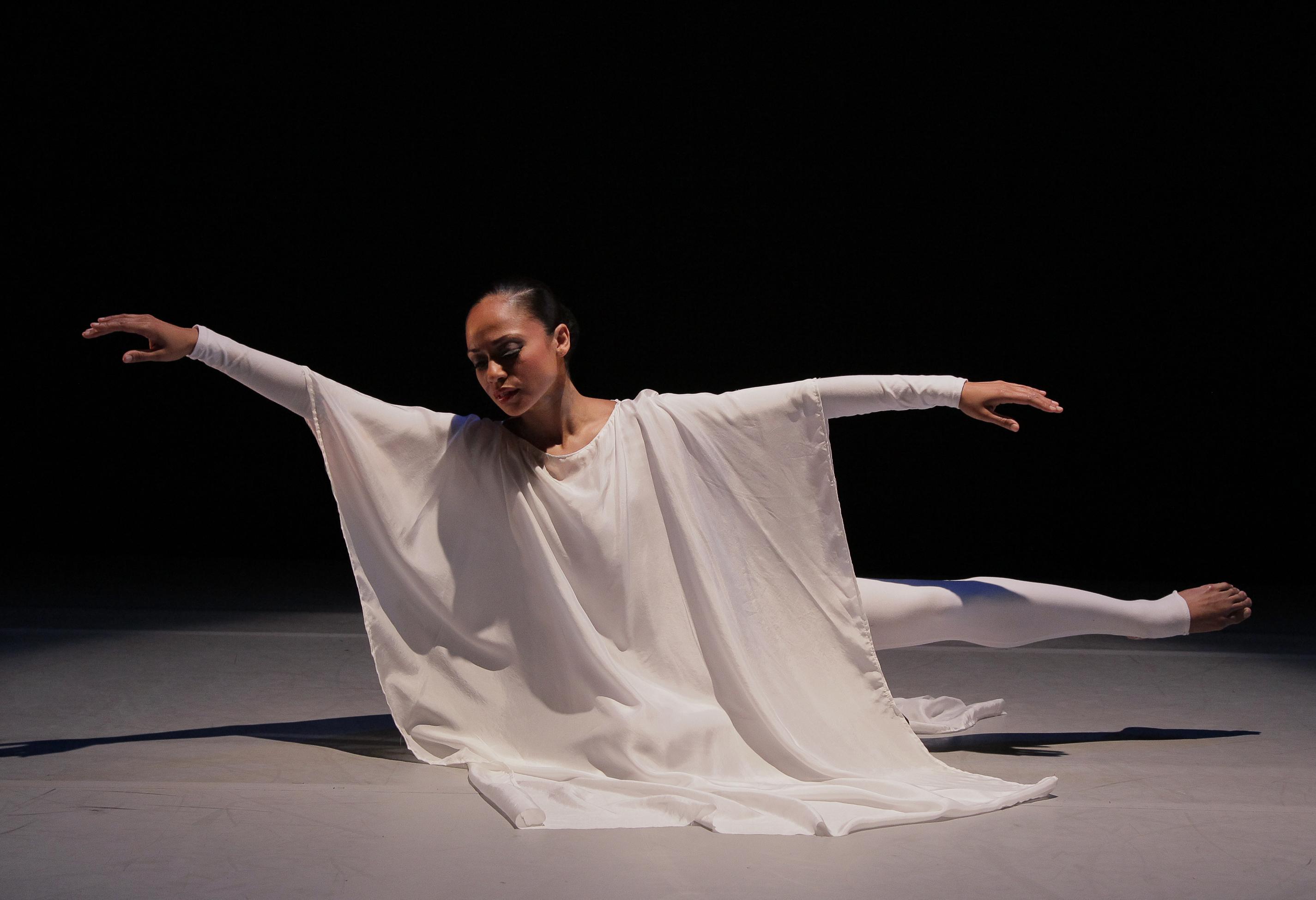 A dancer in a flowing white costume performing on stage, leaning forward with one leg extended behind and arms gracefully outstretched. The background is dark, emphasizing the elegance and poise of the dancer's pose. The lighting casts soft shadows, enhancing the ethereal quality of the performance.