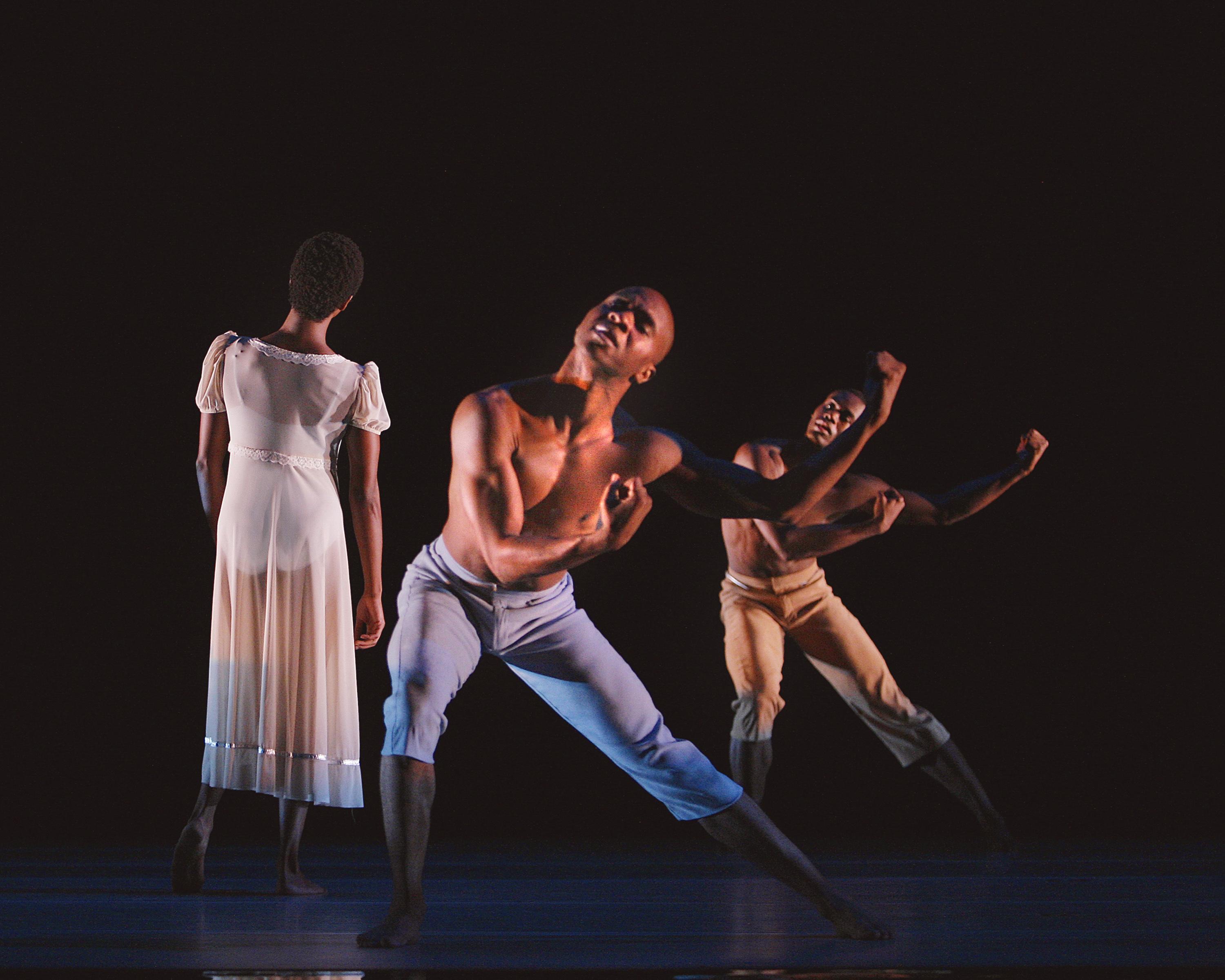 Three dancers on stage. A woman in a long, flowing white dress stands with her back to the camera. Two shirtless men in beige and light blue pants are in dynamic poses, leaning forward with their arms bent. The background is dark.