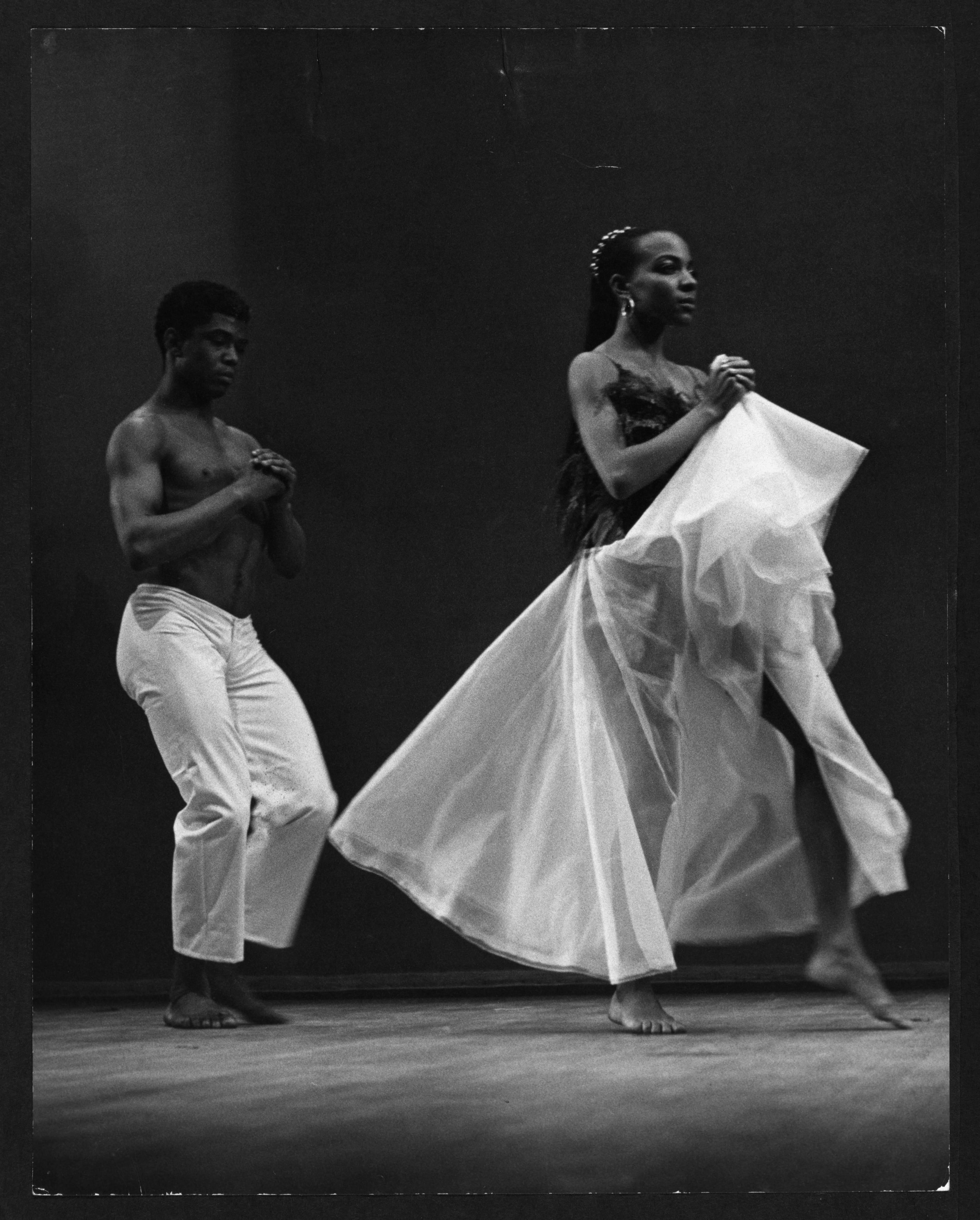 A black and white photo of two dancers. A shirtless male dancer in white pants stands with hands clasped. A female dancer in a flowing white dress moves with hands clasped. The background is dark.