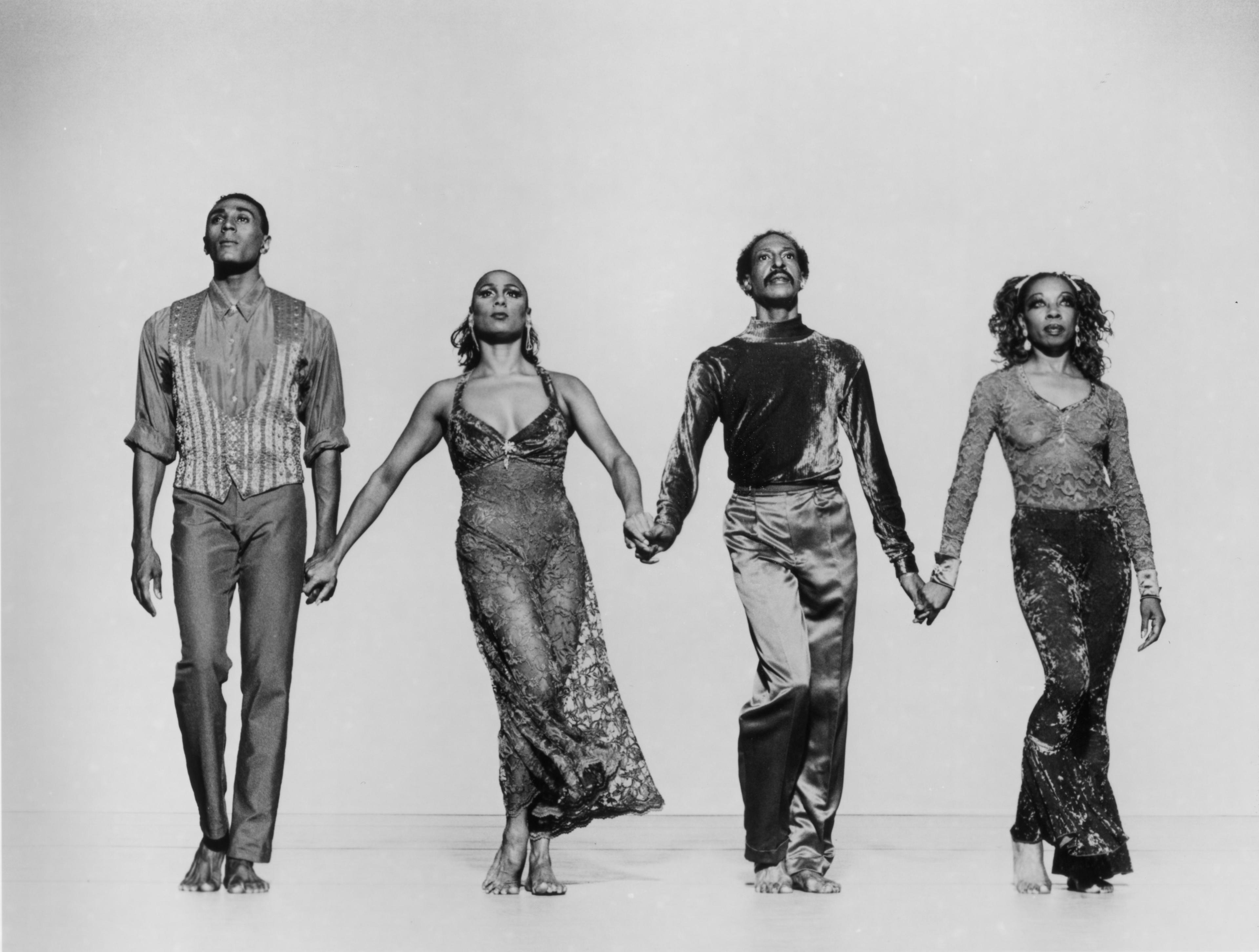 A black and white photo of four dancers holding hands, walking forward. Two male dancers and two female dancers are wearing patterned costumes. The background is light.