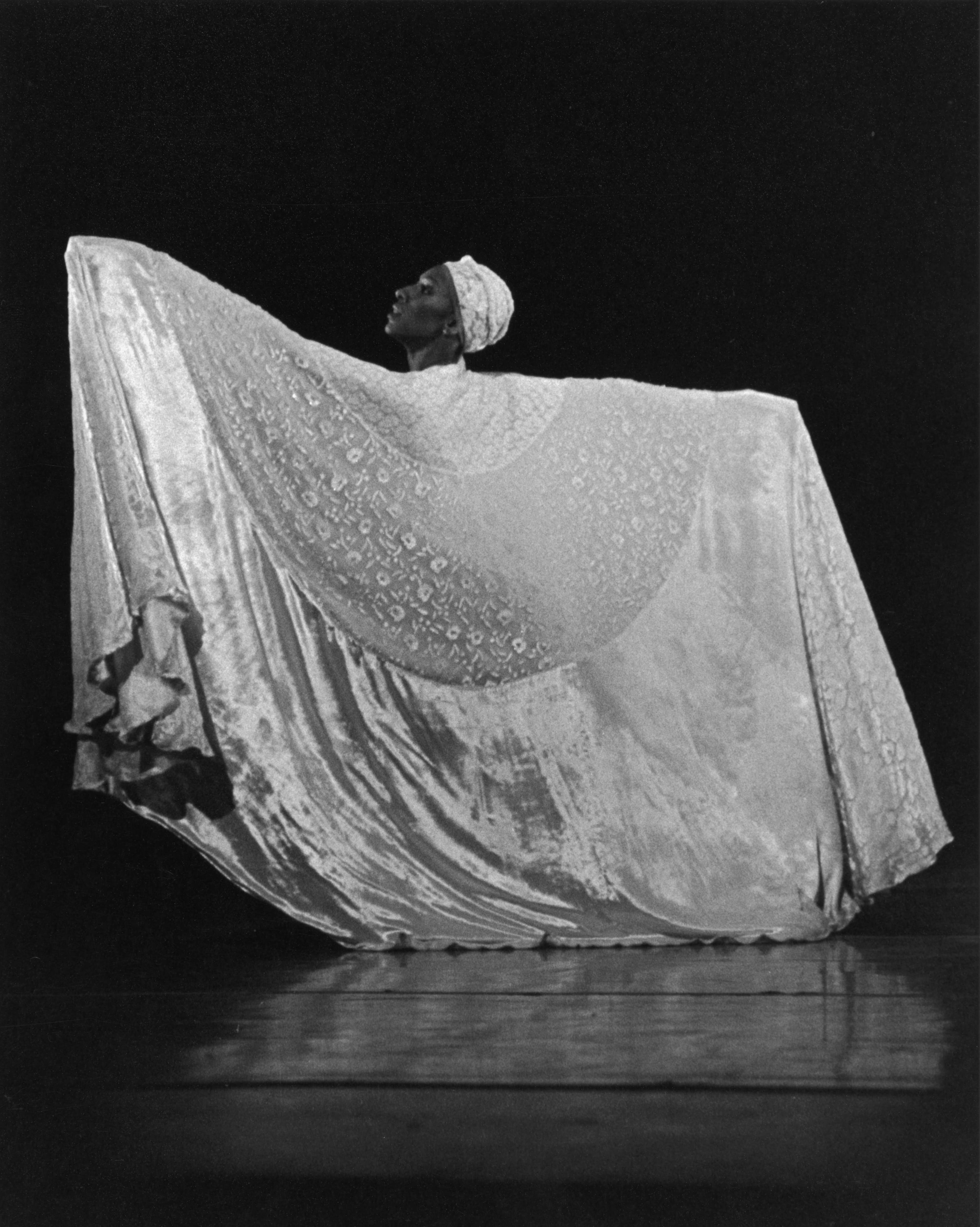 A black and white photo of a female dancer wearing a large, flowing garment and a headwrap. She stands with arms extended, holding the fabric wide. The background is dark.