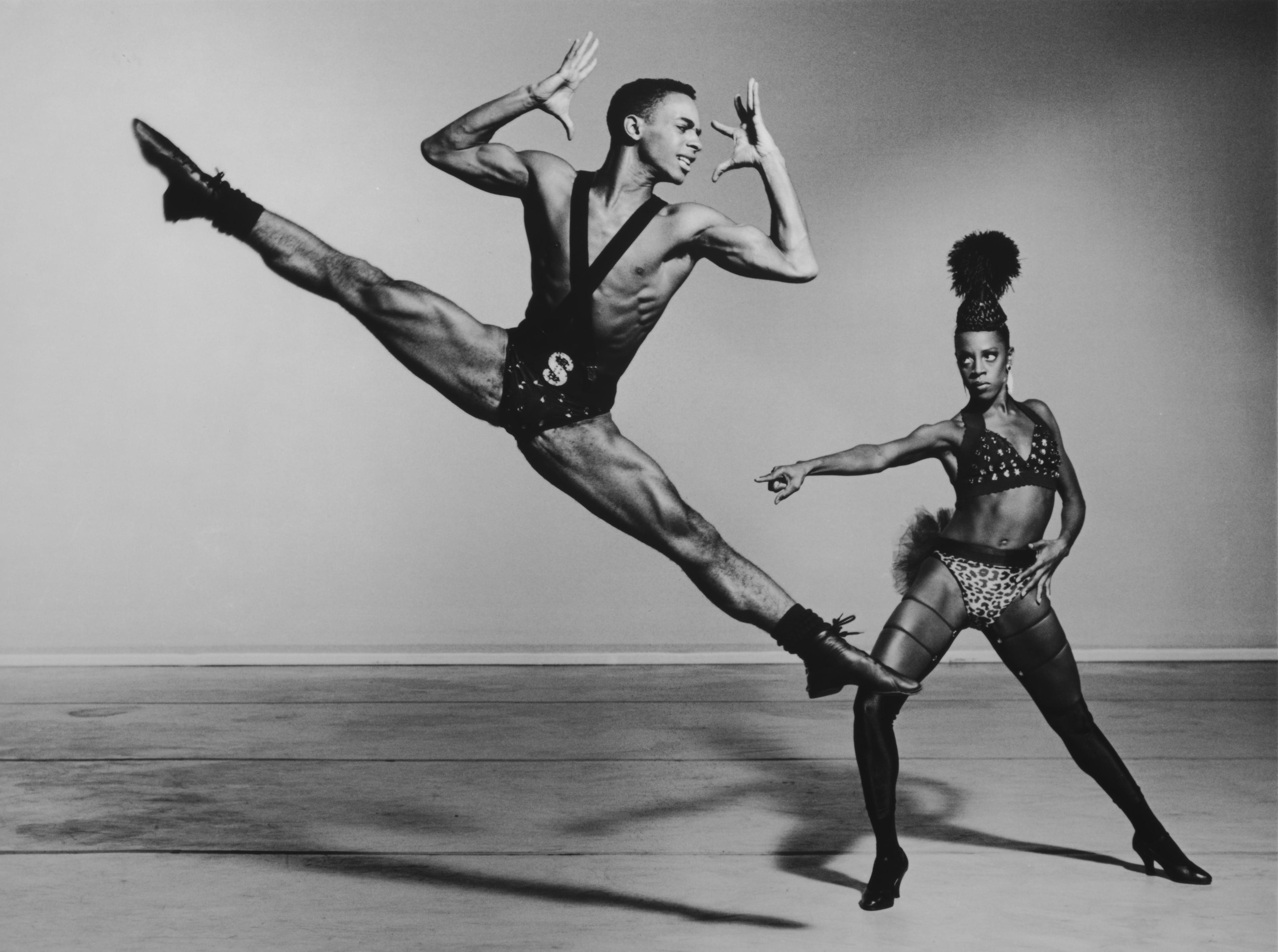 A black and white photo of two dancers. A male dancer wearing a harness and briefs with an "S" leaps in the air. A female dancer in a studded top and leopard print briefs, with a tall headpiece, points and looks at him. The background is light.