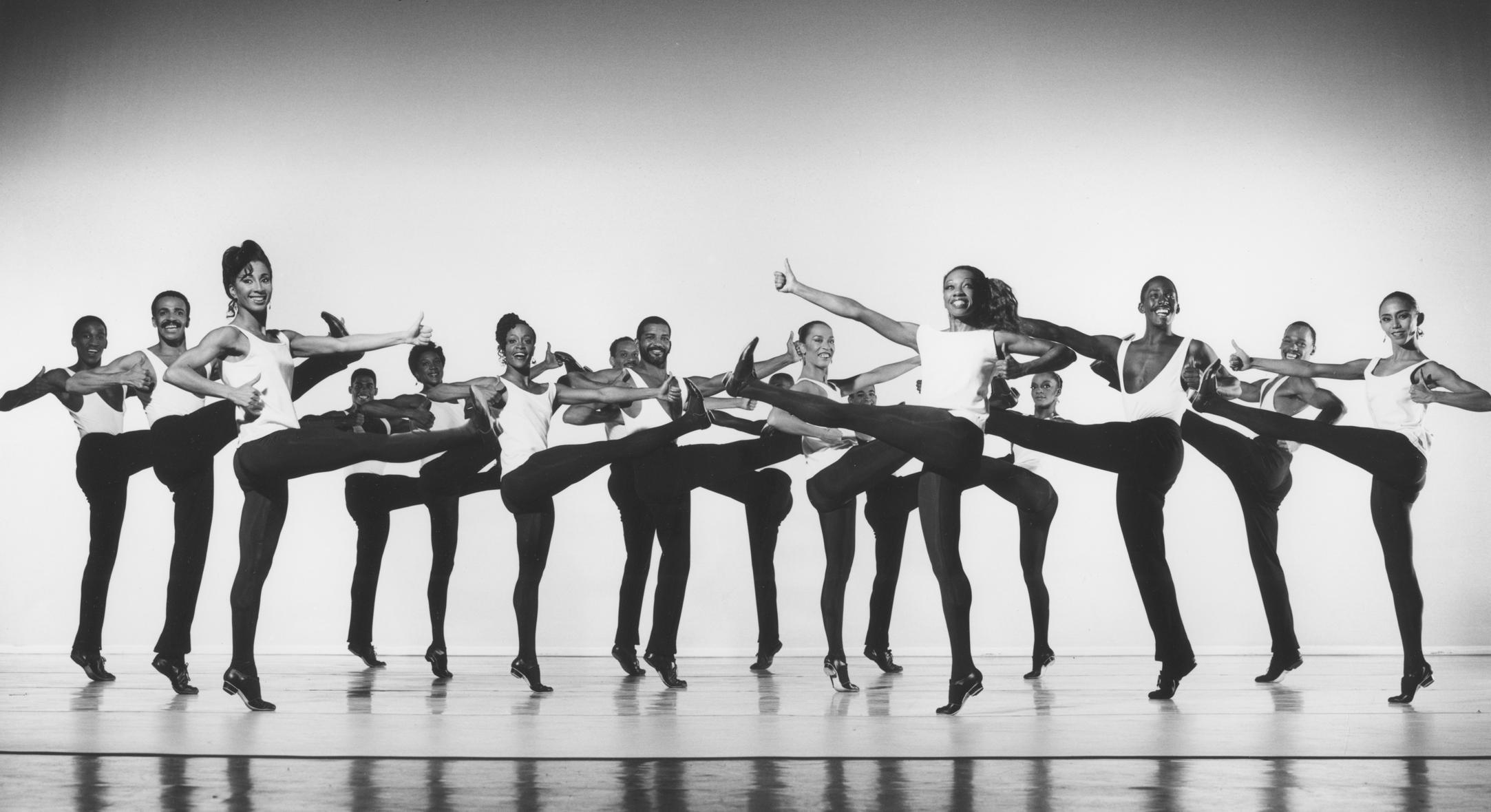 A black and white photo of a group of dancers performing in unison. They are all wearing white tops and black pants, and are mid-kick with one leg raised and arms extended. The background is light.