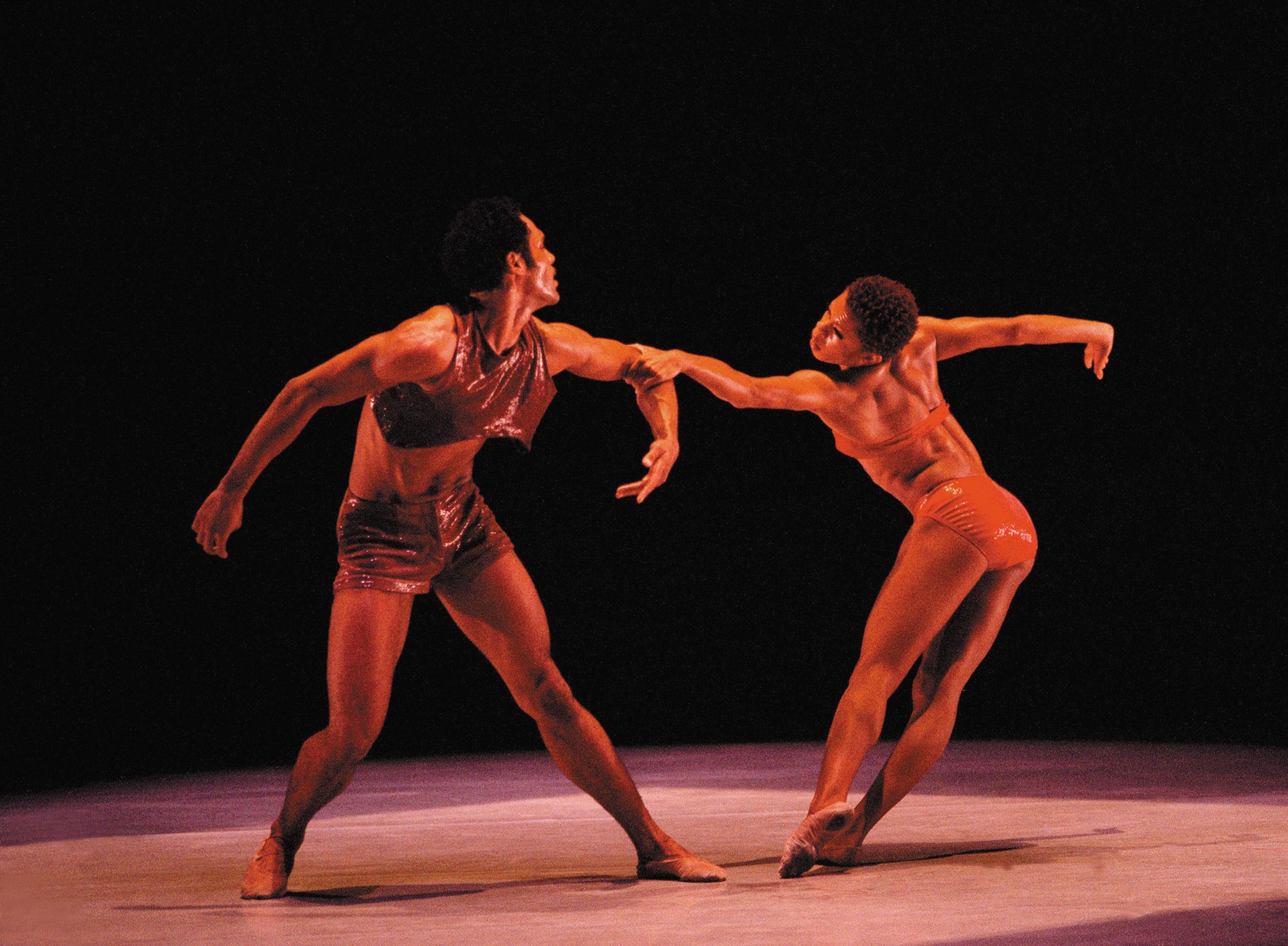 Two dancers on stage in dynamic poses. The male dancer wears a sleeveless top and shorts in a shiny red fabric. The female dancer wears a red two-piece costume. They are reaching out towards each other with extended arms. The background is dark.