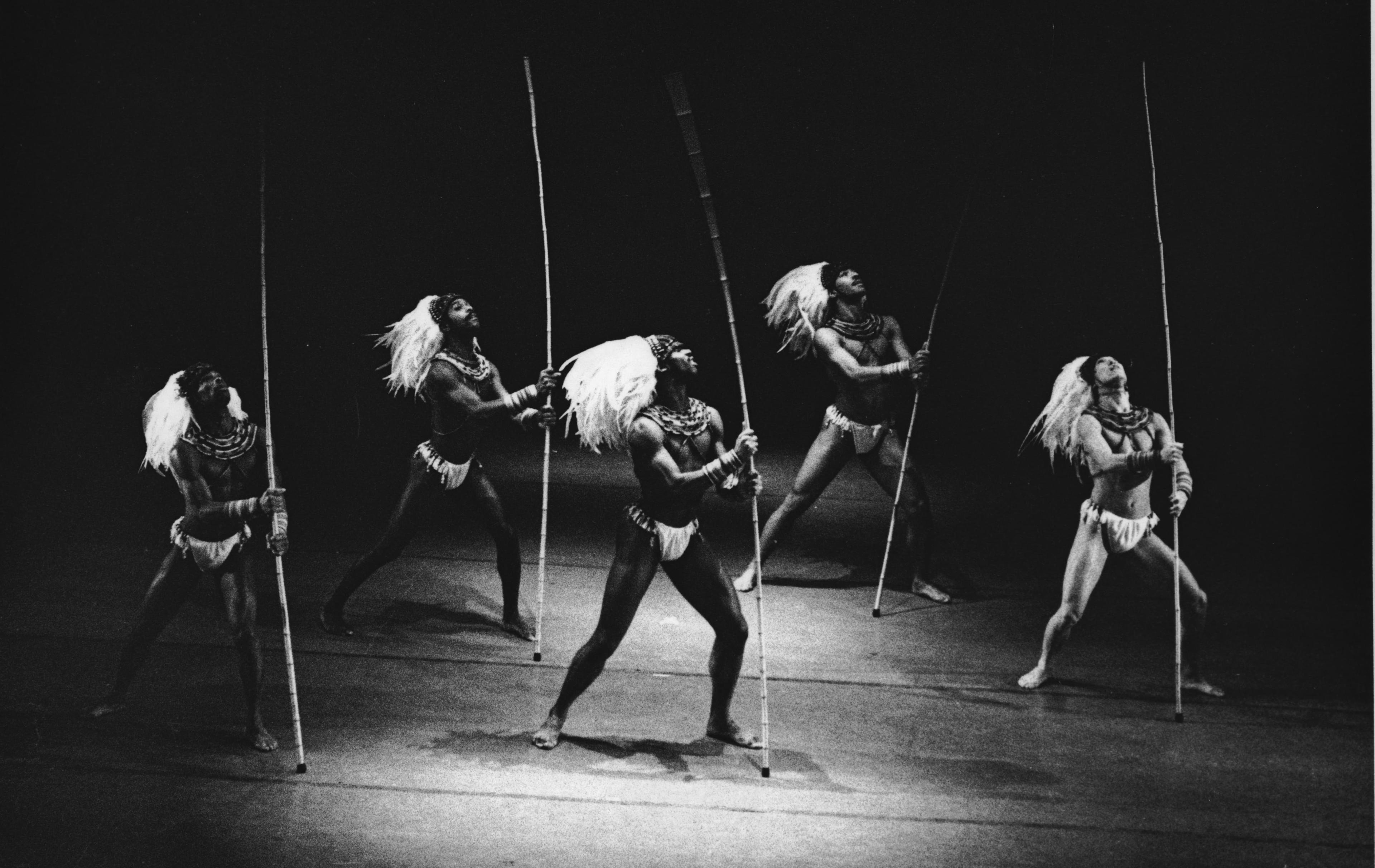 A black and white photo of five male dancers on stage, each holding a long pole. They are dressed in tribal-inspired costumes with feathered headdresses and necklaces. They are in various dynamic poses, looking upward. The background is dark.