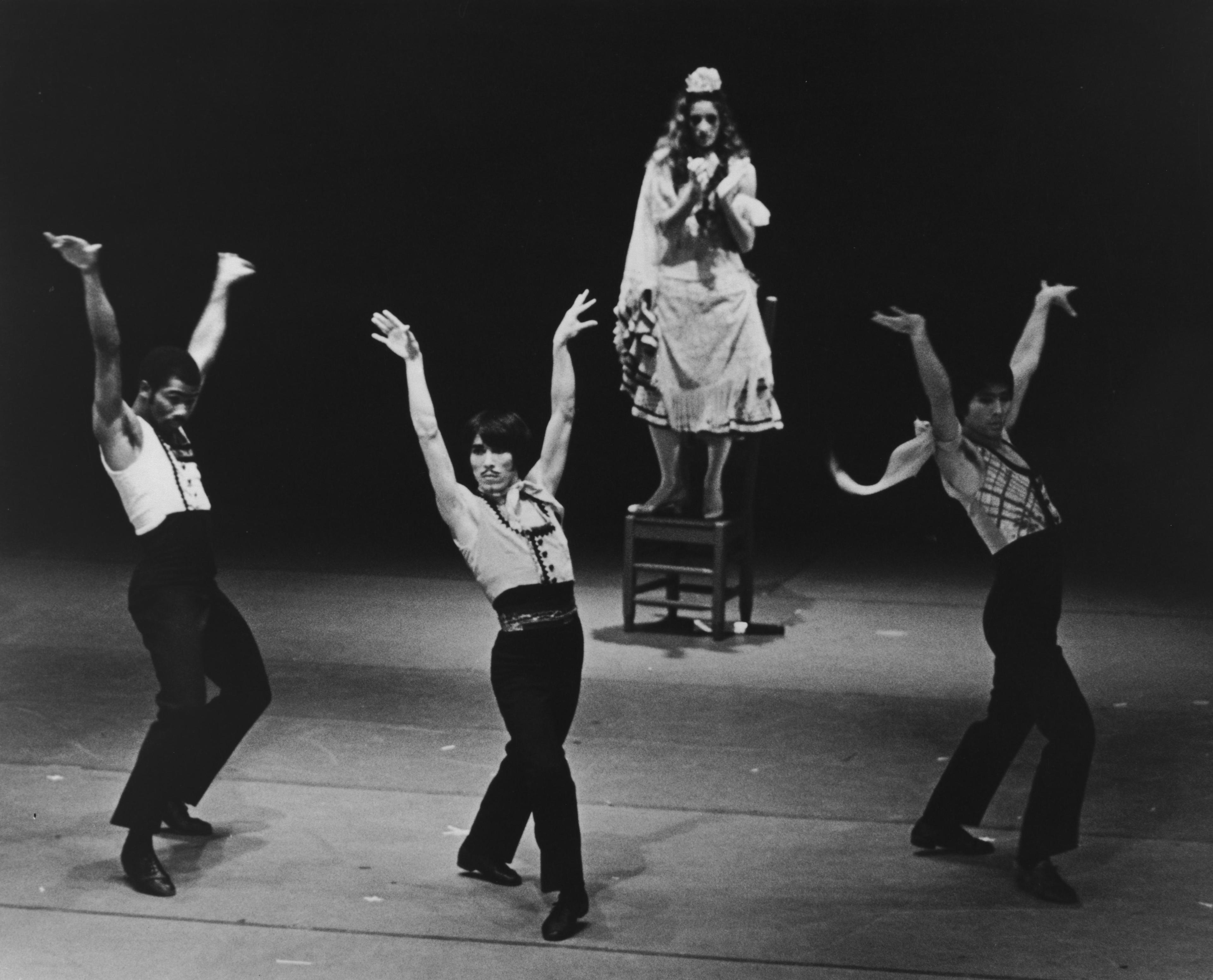 A black and white photo of three male dancers in matching costumes performing with their arms raised. A female dancer stands on a chair in the background, wearing a dress and headscarf. The background is dark.