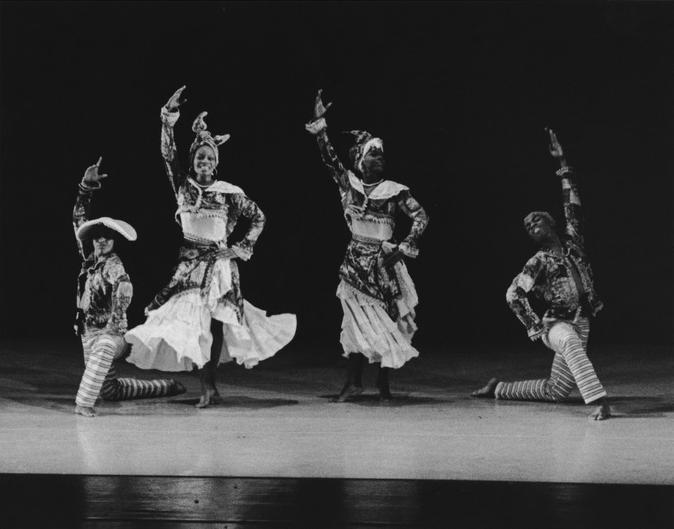 Four dancers on stage in elaborate costumes. Two dancers are standing with one leg raised and arms extended upward. The other two dancers are kneeling with one arm raised. They are dressed in patterned dresses, striped stockings, and headwraps. The background is dark.