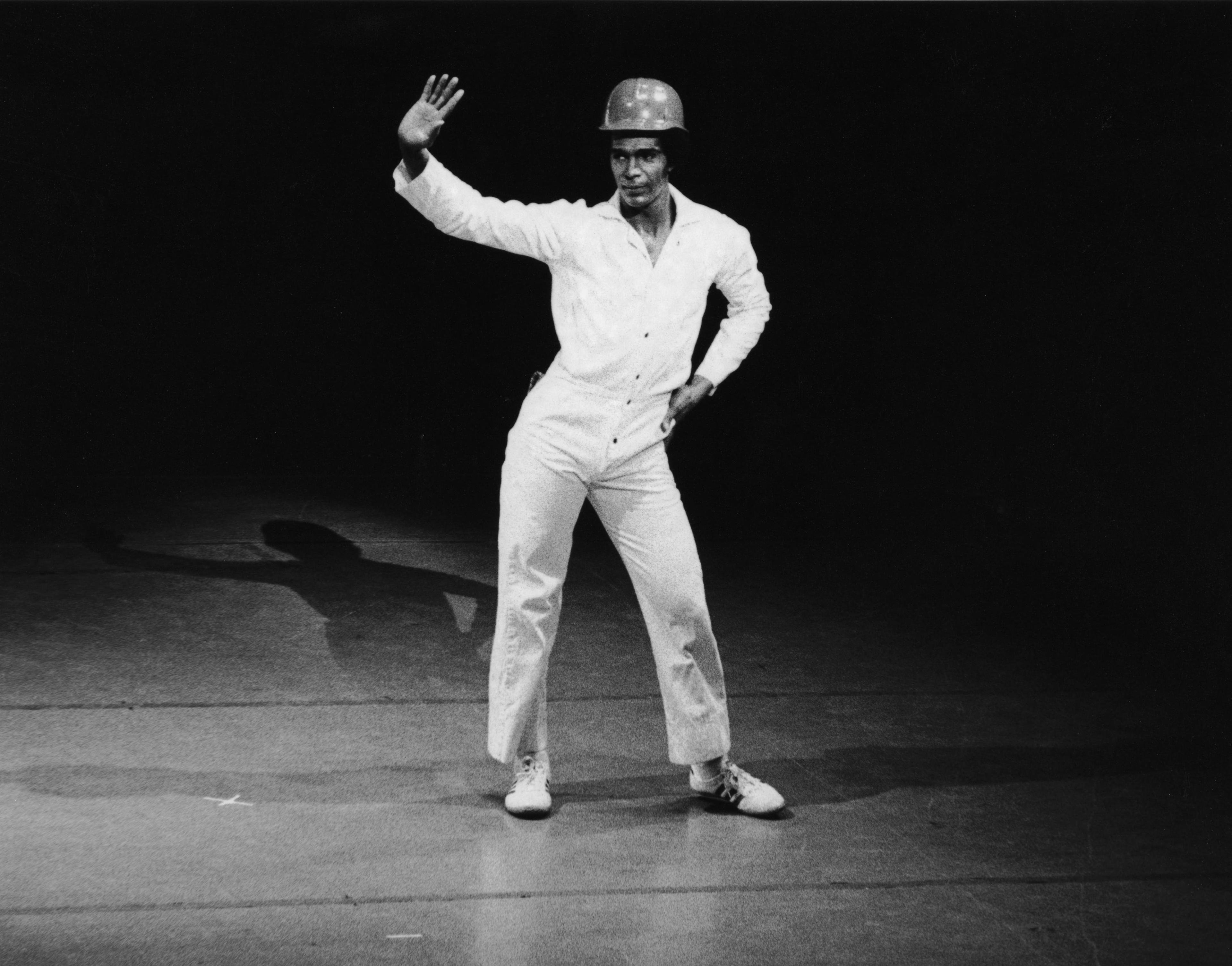 A black and white photo of a male dancer wearing a white jumpsuit and a hard hat. He is standing with one hand on his hip and the other raised, striking a confident pose. The background is dark.