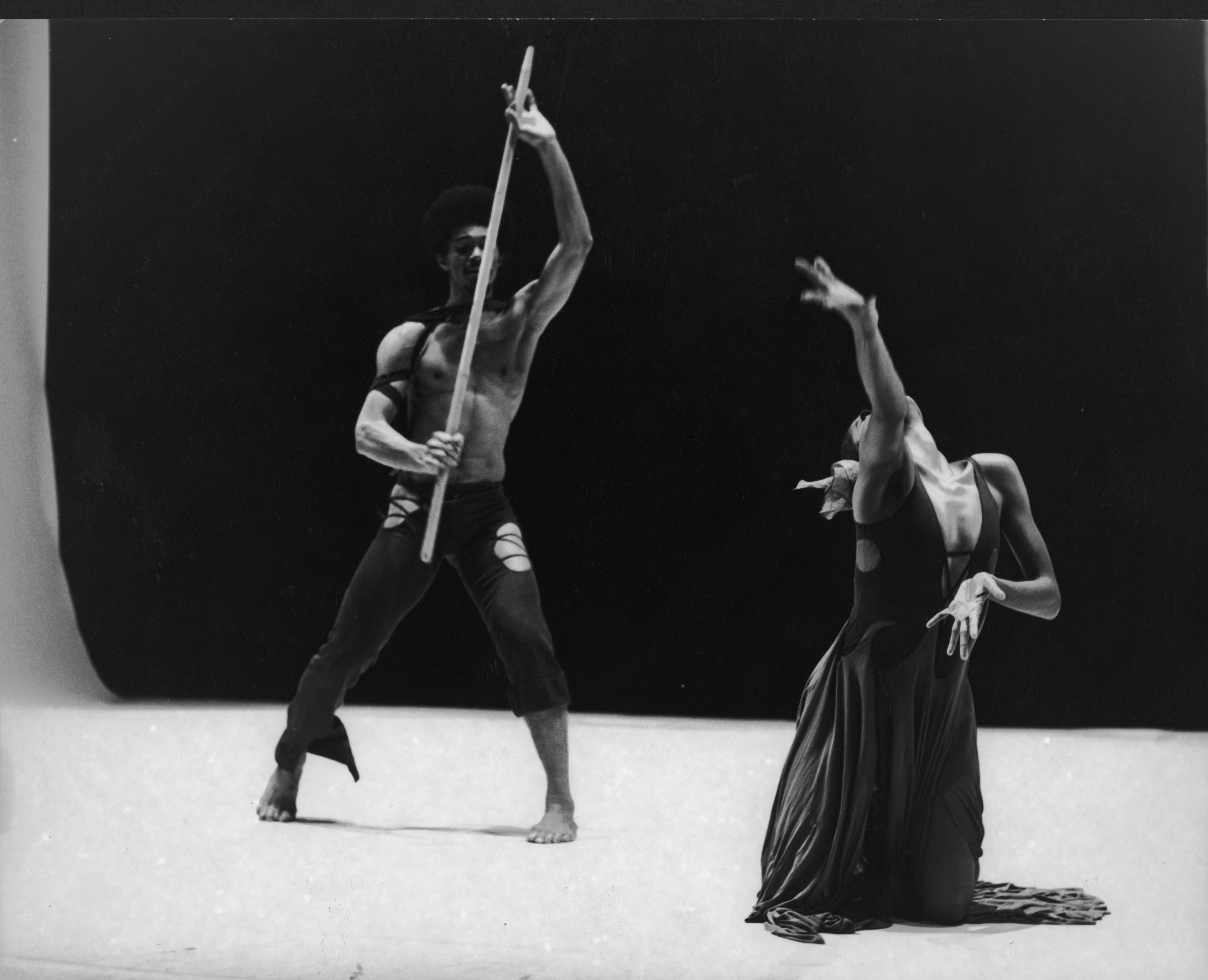 A black and white photo of two dancers, Consuelo Atlas and Clive Thompson, in mid-performance. Clive Thompson stands with a raised wooden stick, wearing dark, torn pants. Consuelo Atlas is kneeling and leaning back dramatically, wearing a dark flowing dress. The background is dark, highlighting the dancers. The photo is labeled REPERTORY Consuelo Atlas and Clive Thompson in Child of the Earth.