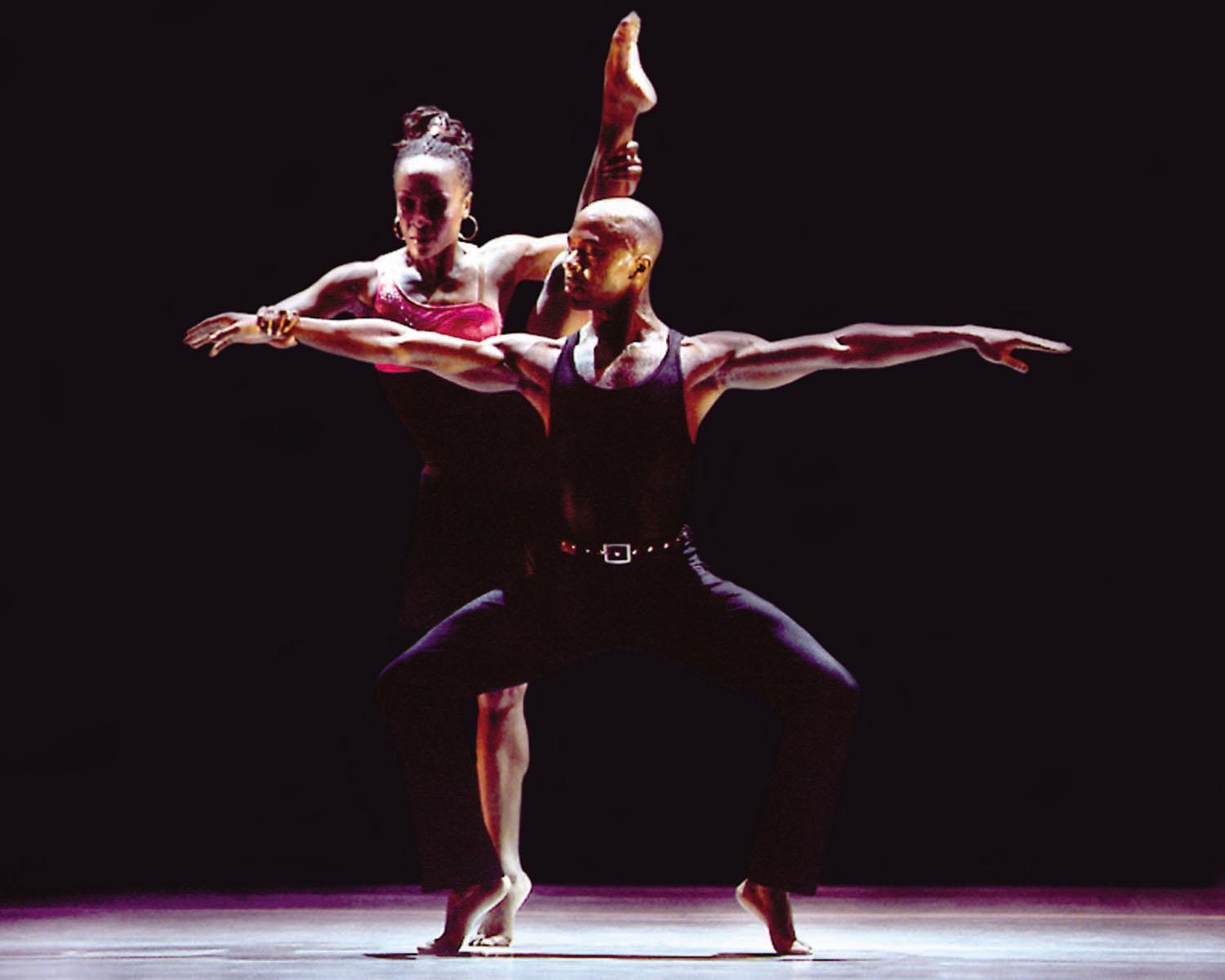 A color photo of two dancers in mid-performance. The male dancer in the foreground is in a deep lunge with arms extended, wearing a black tank top and pants. Behind him, a female dancer is balancing on one leg with the other leg raised high, wearing a red top and black pants. The background is dark, highlighting the dancers. 