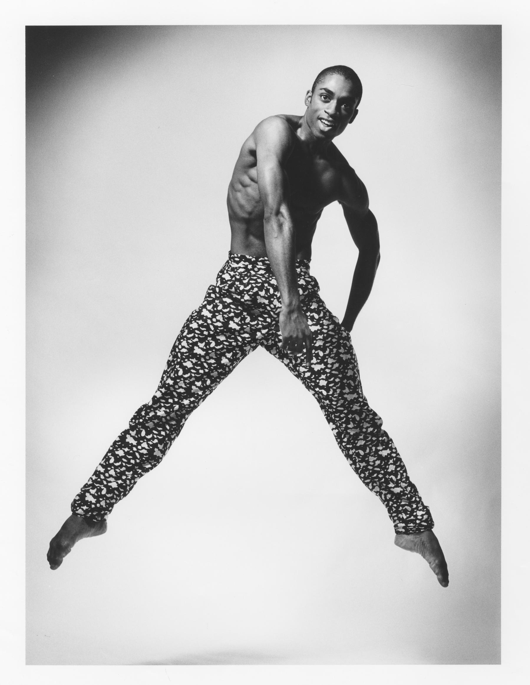 A black and white photo of dancer Desmond Richardson in mid-air. He is shirtless, wearing patterned pants with a black and white design. He has one arm bent at his side and the other extended downward, with legs apart. The background is plain and light-colored. The photo is labeled REPERTORY Desmond Richardson in Escargot. Photo by Jack Mitchell.