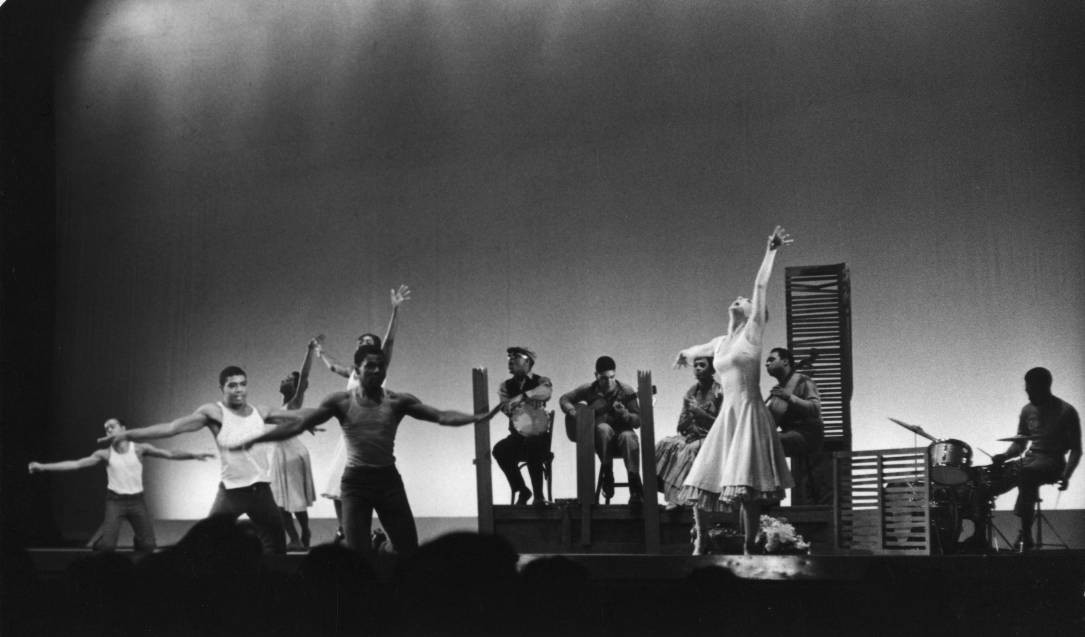 A black and white photo of a performance featuring multiple dancers and musicians. Dancers are in various poses across the stage, some with arms extended. Musicians sit in the background, playing instruments like the guitar and drums. The stage is set with minimal props. The photo is labeled REPERTORY Donald Martin, Alvin Ailey, Minnie Marshall, Georgia Collins, Ella Thompson, Charles Moore, Carmen de Lavallade, Brother John Sellers, Bruce Langhorn, Horace Arnold in Been Here and Gone. 