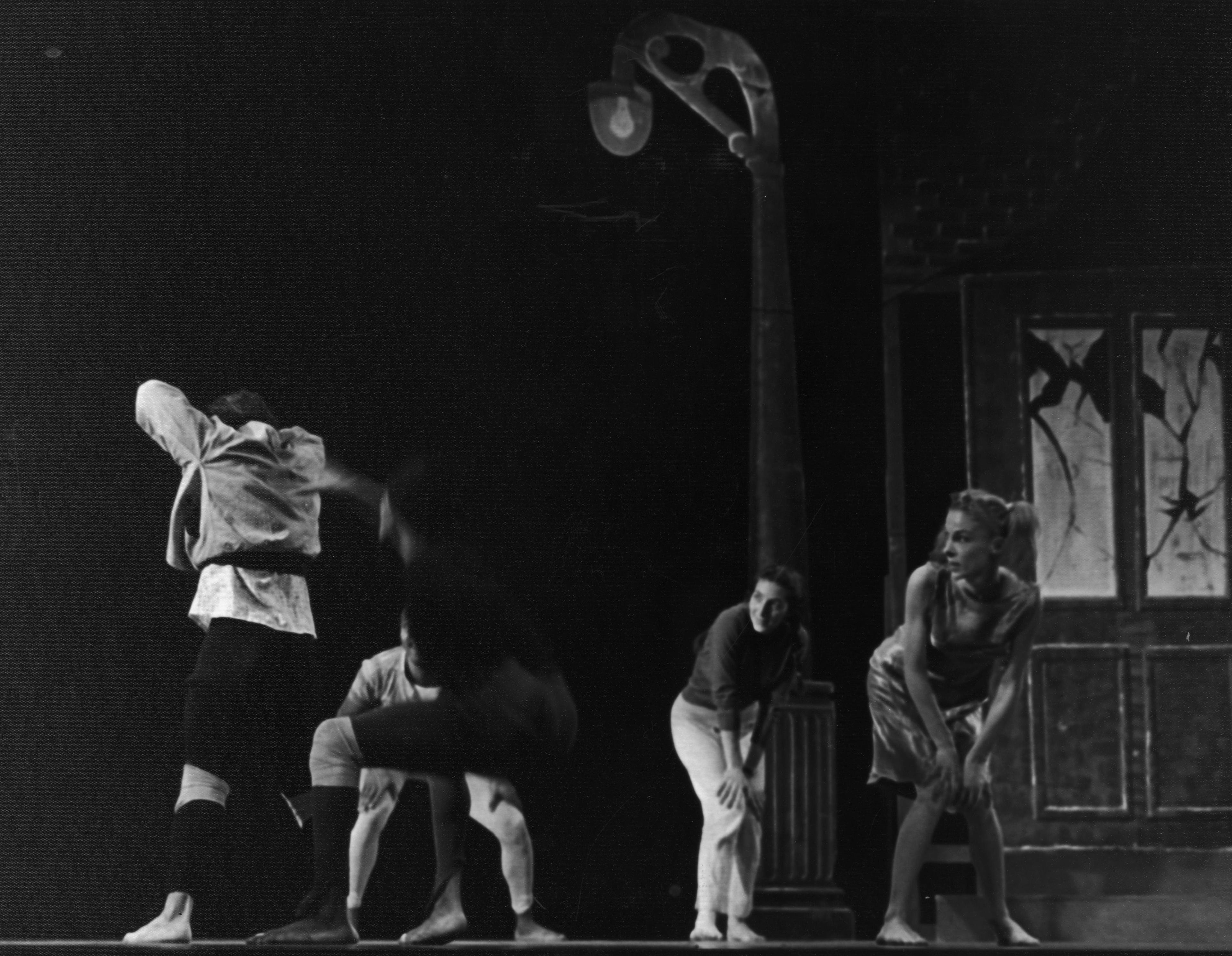 A black and white photo of a performance by Donald McKayle & Co. in Games. Several dancers are on stage, some crouching and others mid-movement. They are barefoot and wearing casual costumes. A streetlamp and a building with broken windows serve as the backdrop.