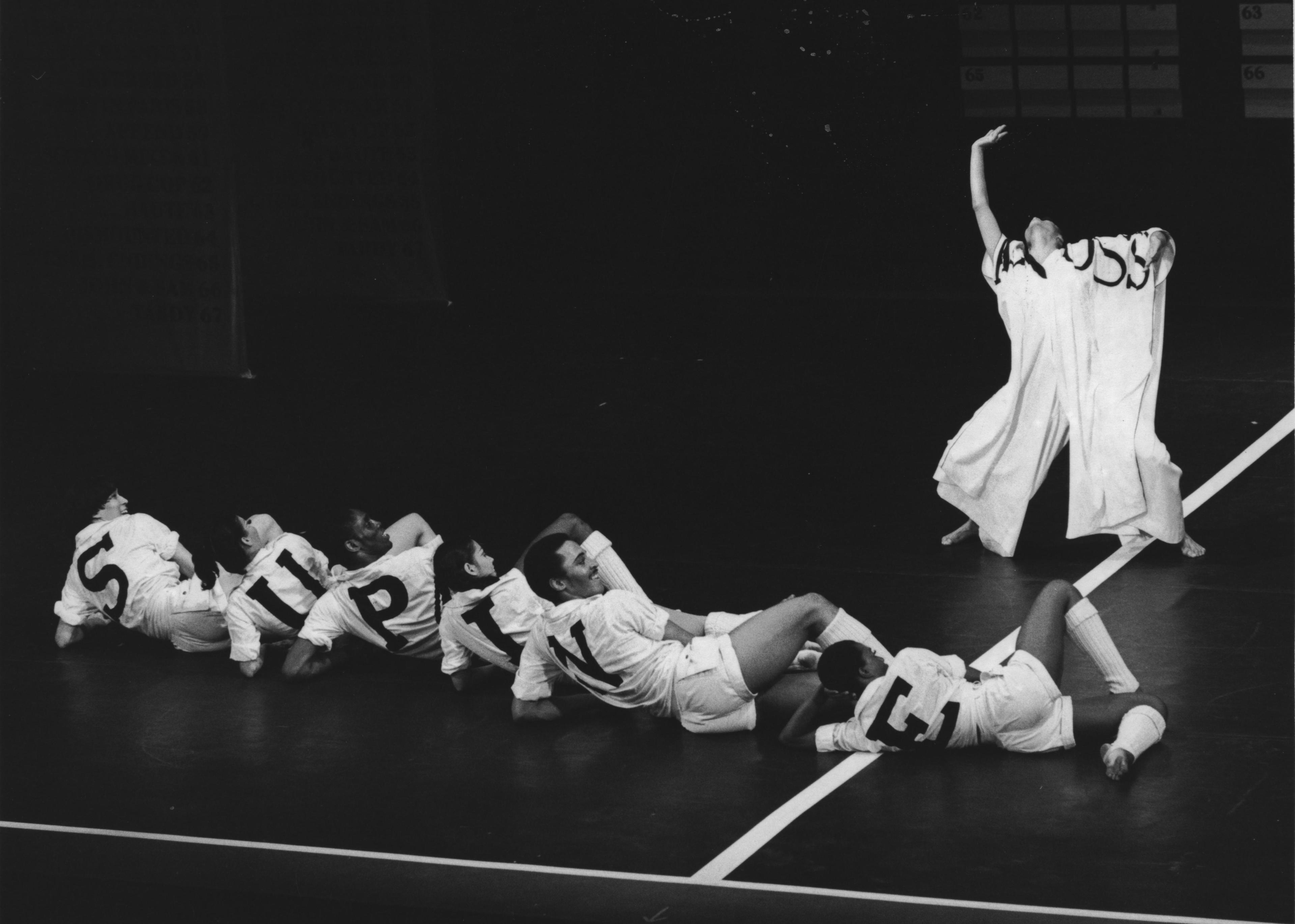 A black and white photo of a performance by Donna Wood and the Company in Crossword. Several dancers are lying on the floor in a row, each wearing white costumes with letters that spell out SPRING. One dancer stands to the side with an arm raised, wearing a flowing white garment. The background is dark, highlighting the dancers.