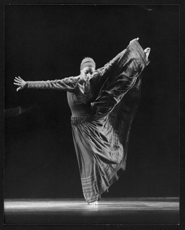 A black and white photo of dancer Donna Wood in mid-performance. She is wearing a long, flowing dress and a headscarf. One leg is raised high to the side, and her arms are extended, creating a dynamic and graceful pose. The background is dark, highlighting her figure.