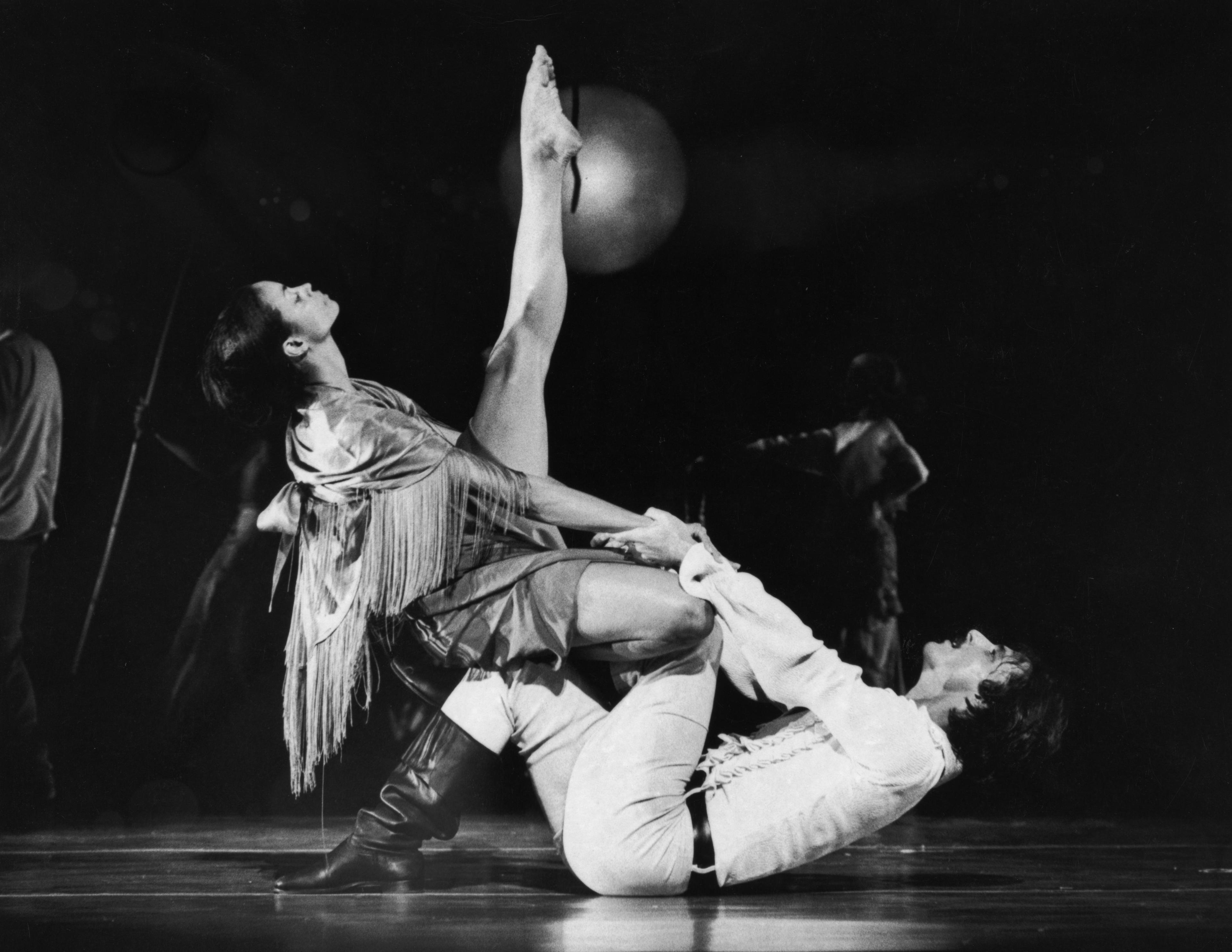 A black and white photo of dancers Donna Wood and Peter Woodin in mid-performance. Donna Wood is sitting on the floor with one leg raised high, supported by Peter Woodin, who is lying on his back and holding her leg. Donna is wearing a fringed garment and boots, while Peter is dressed in light-colored clothes. The background is dark, with other performers visible in the distance. 